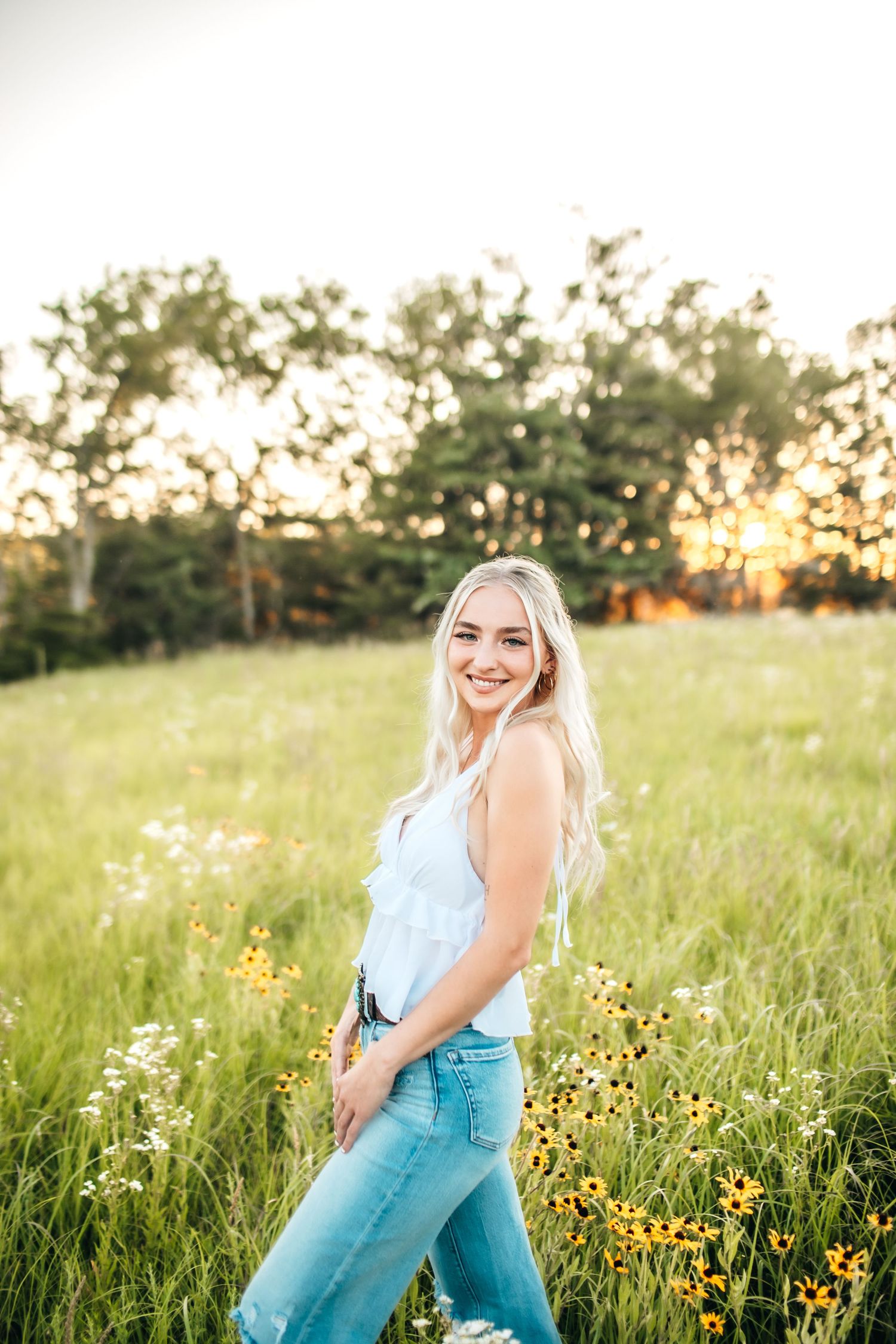 Smiling blonde woman in white top and jeans standing in a sunny wildflower meadow at golden hour.