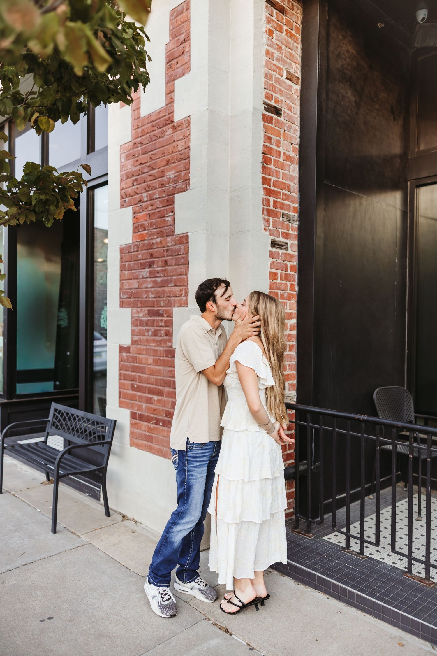 A couple shares a romantic kiss on a city sidewalk beside a brick building, man in jeans and woman in white dress.