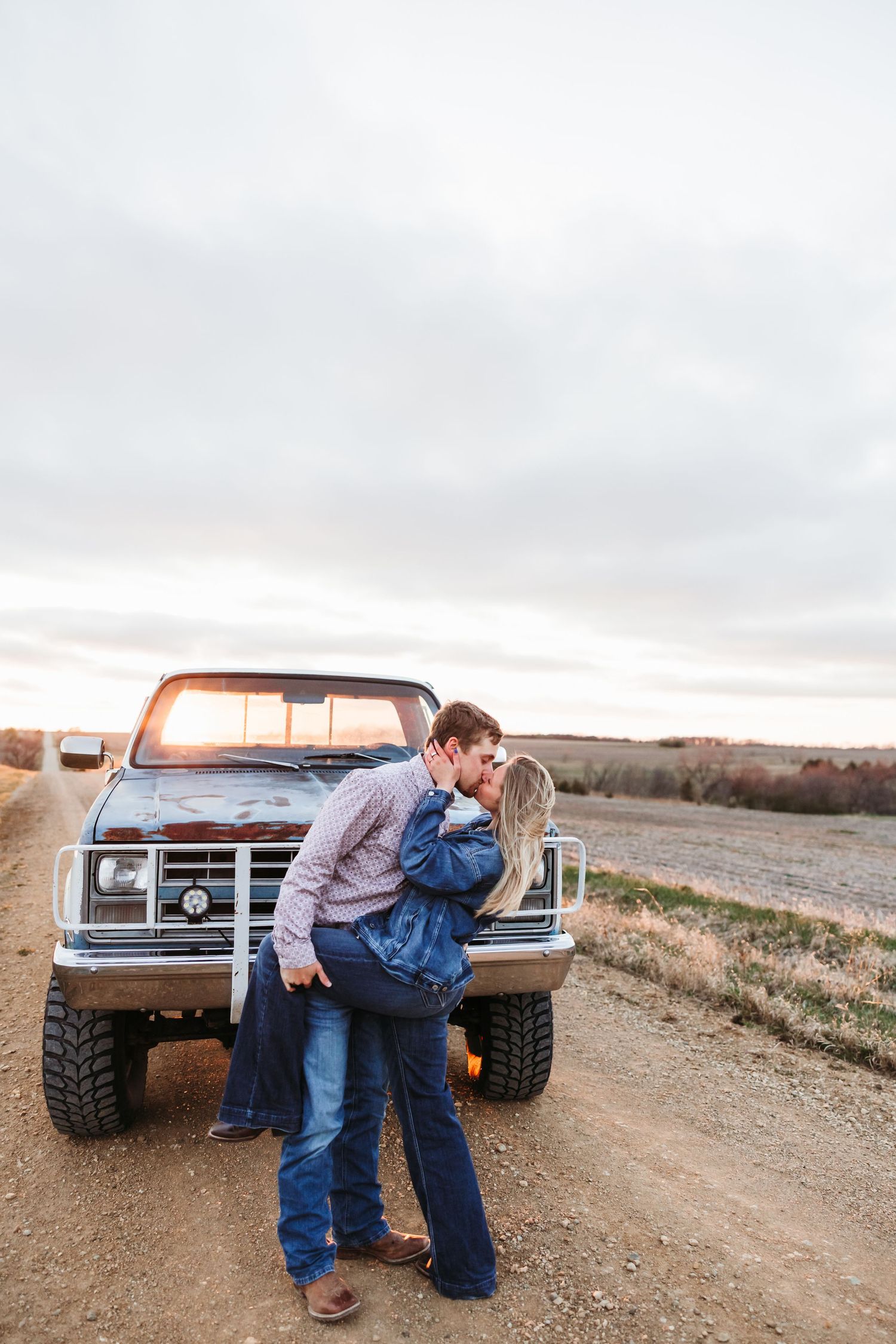 Couple kissing in front of a vintage pickup truck on a rural dirt road at sunset.