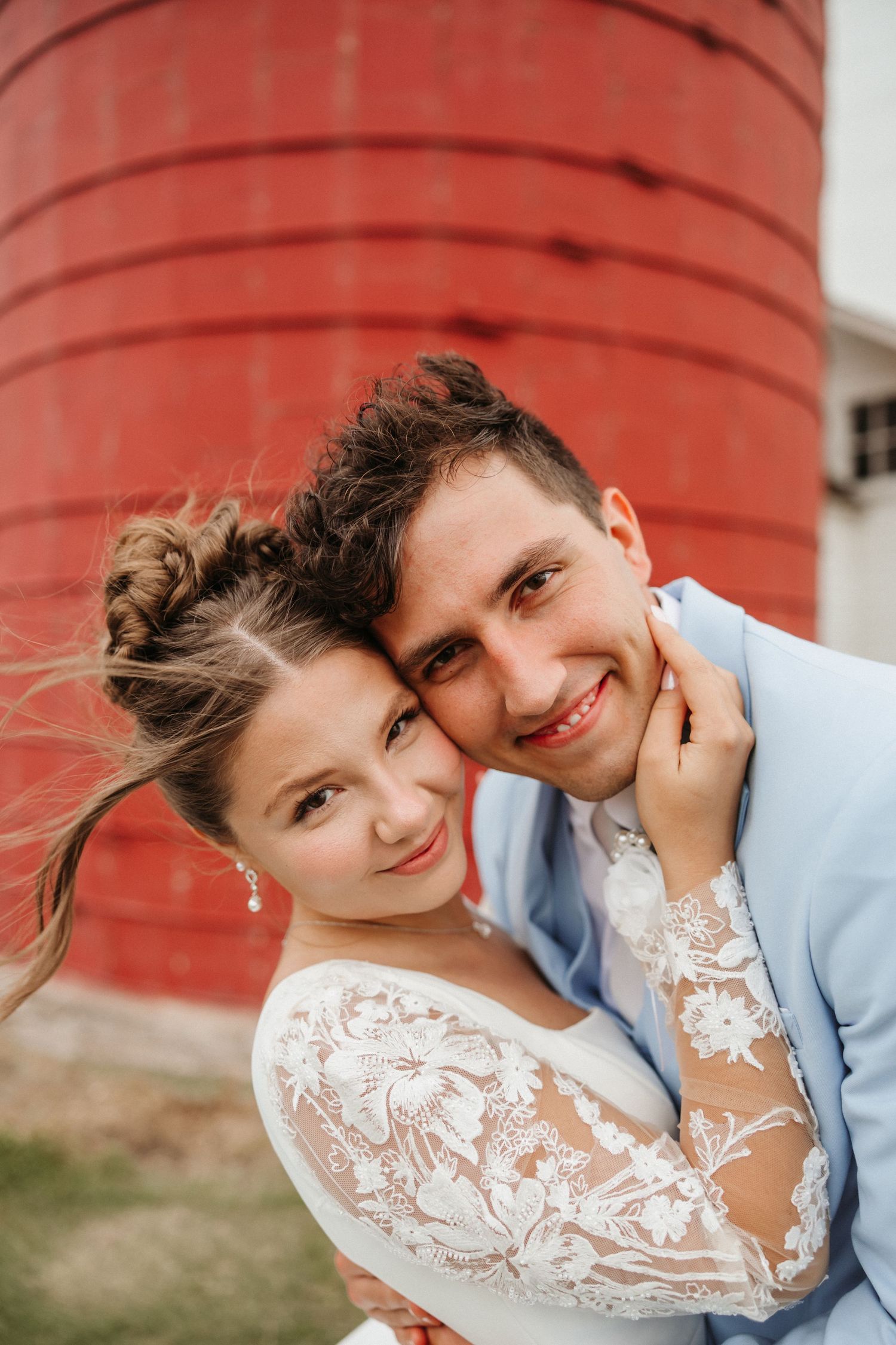 Bride and groom smiling together in front of a red silo, bride in lace dress and groom in light blue suit.