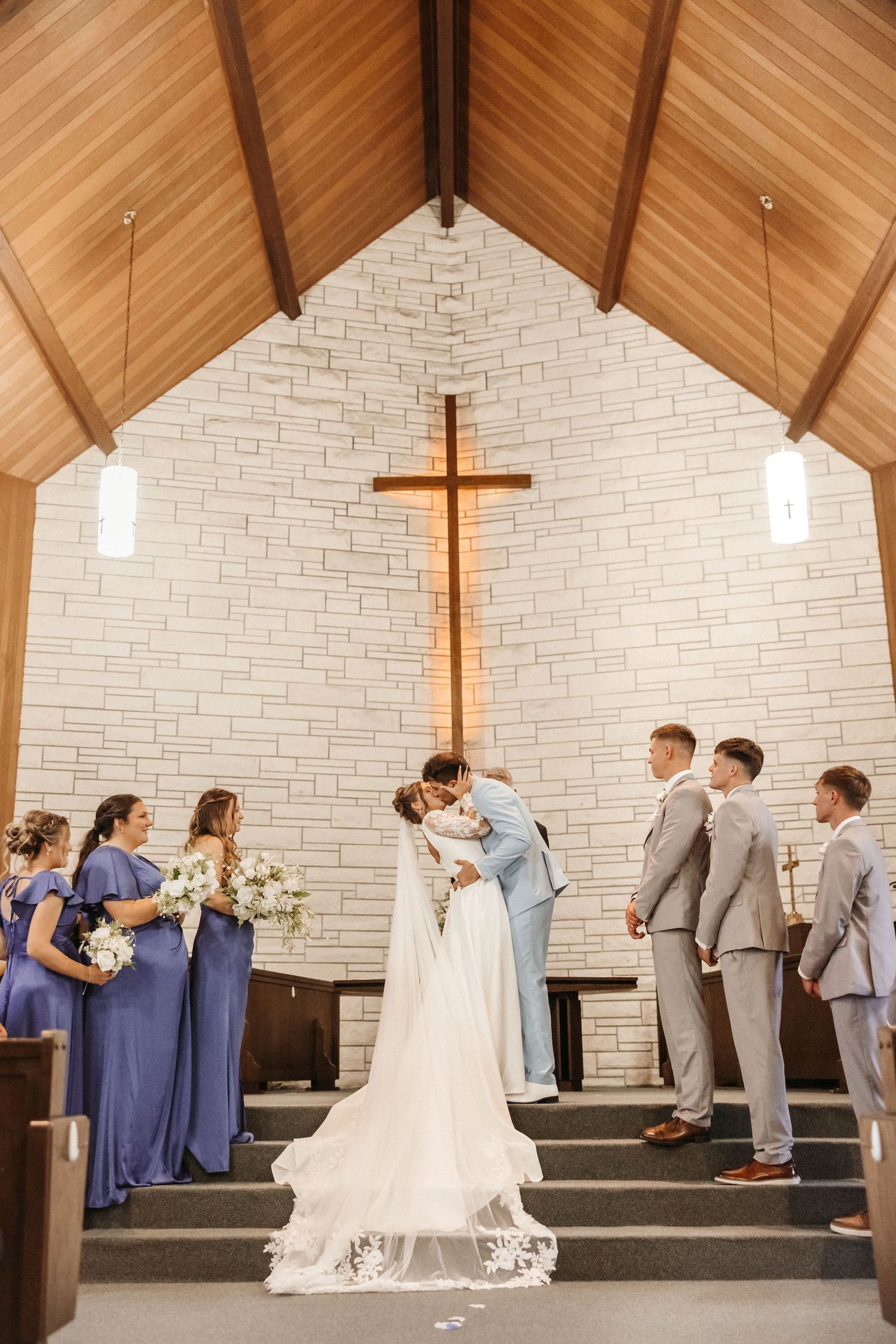 Bride and groom share first kiss at chapel altar, flanked by bridesmaids in blue and groomsmen in gray suits.