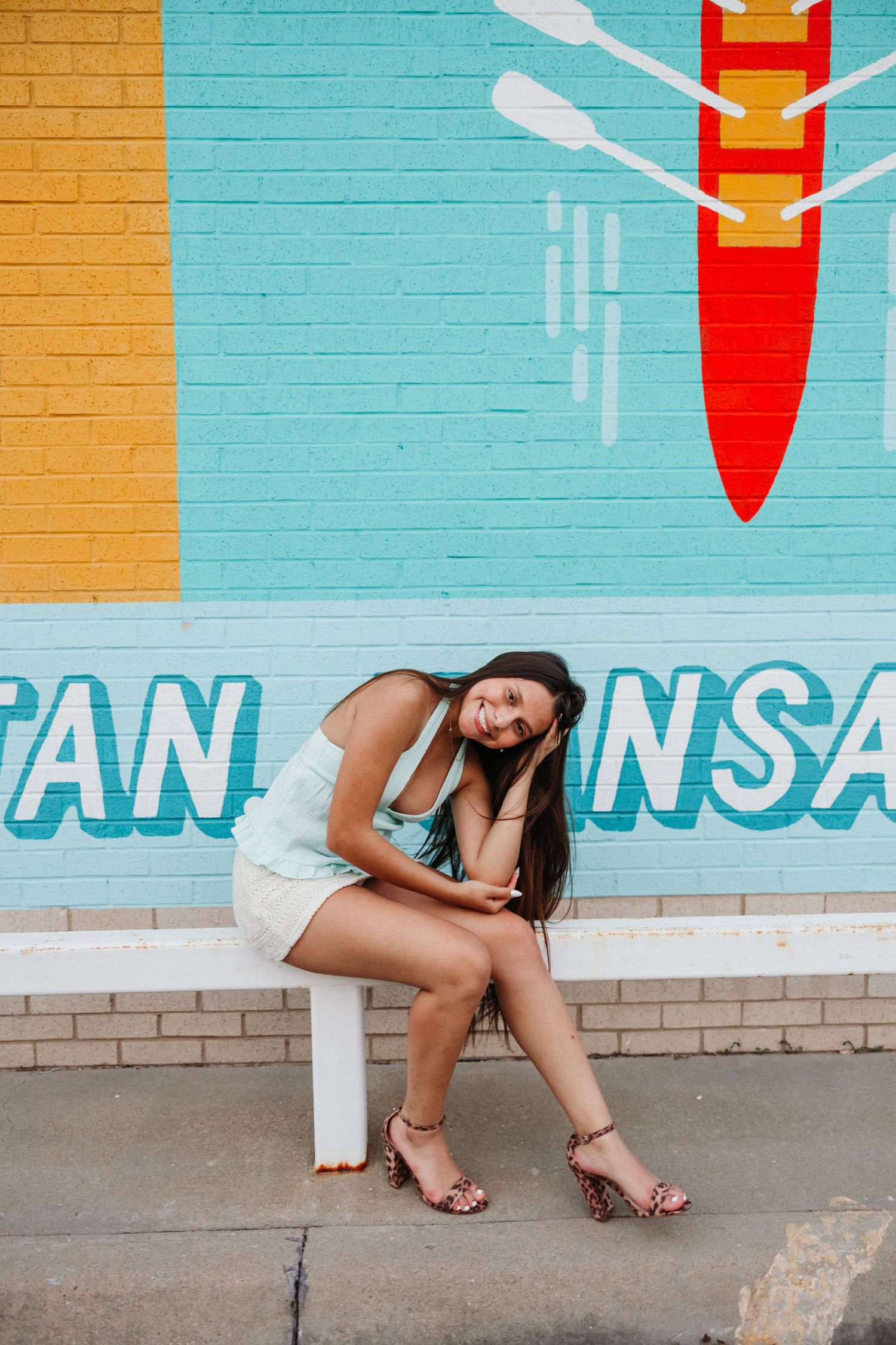 Young woman in white dress and heels sits smiling on a bench in front of a colorful rowing mural.