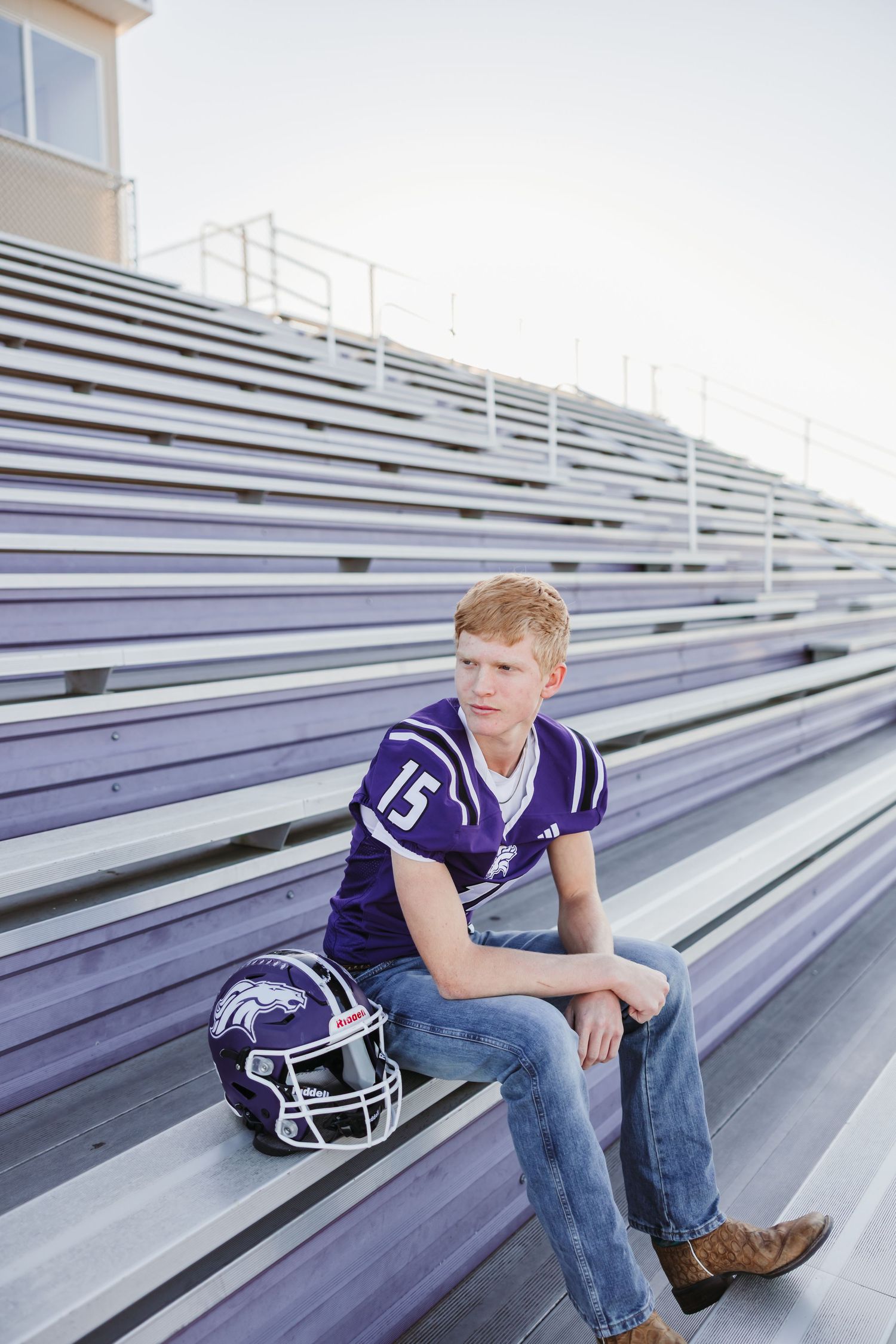 Teen football player in purple jersey #15 sits on bleachers with helmet, wearing jeans and cowboy boots.