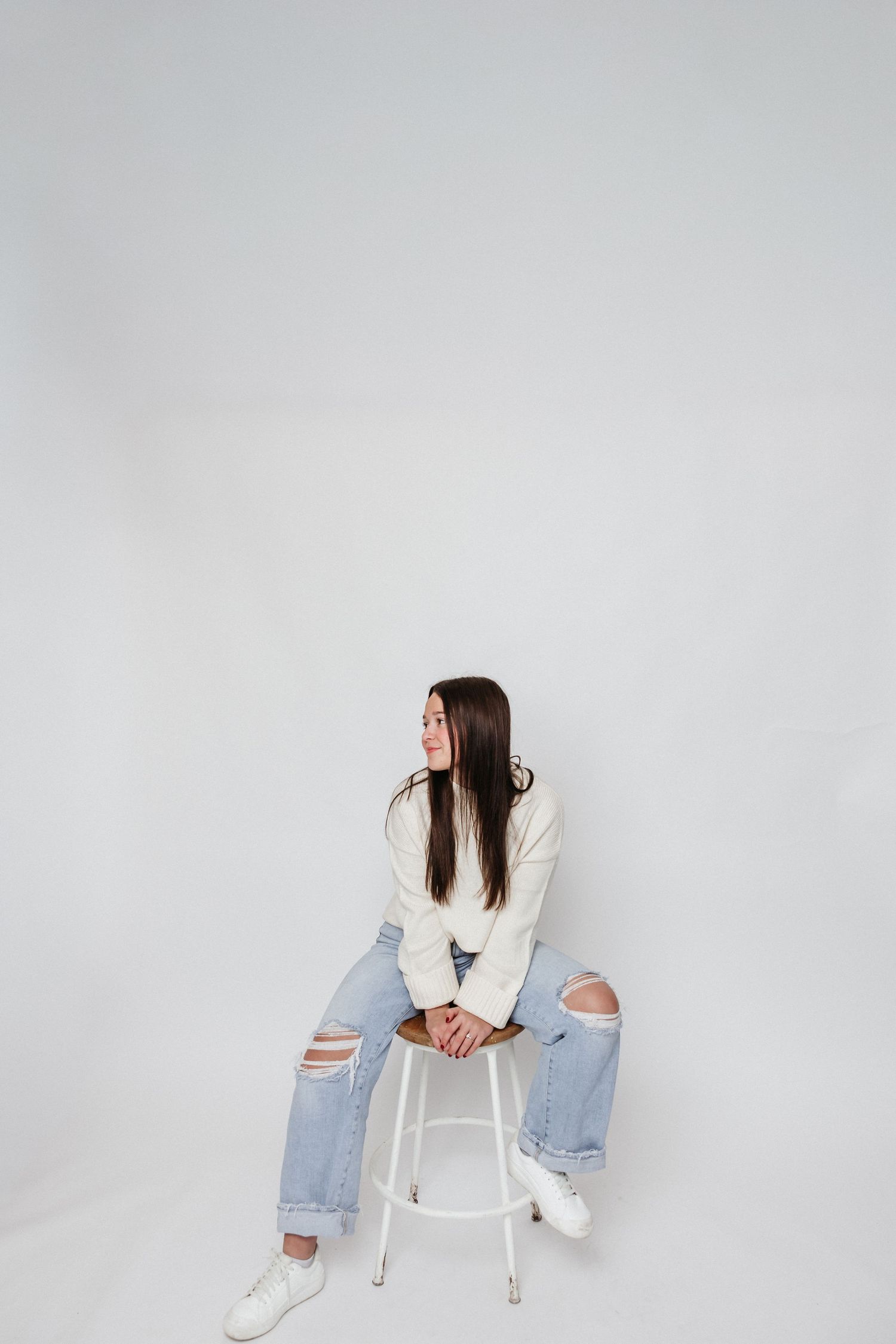 Casual seated pose on white stool wearing distressed denim and cream sweater.