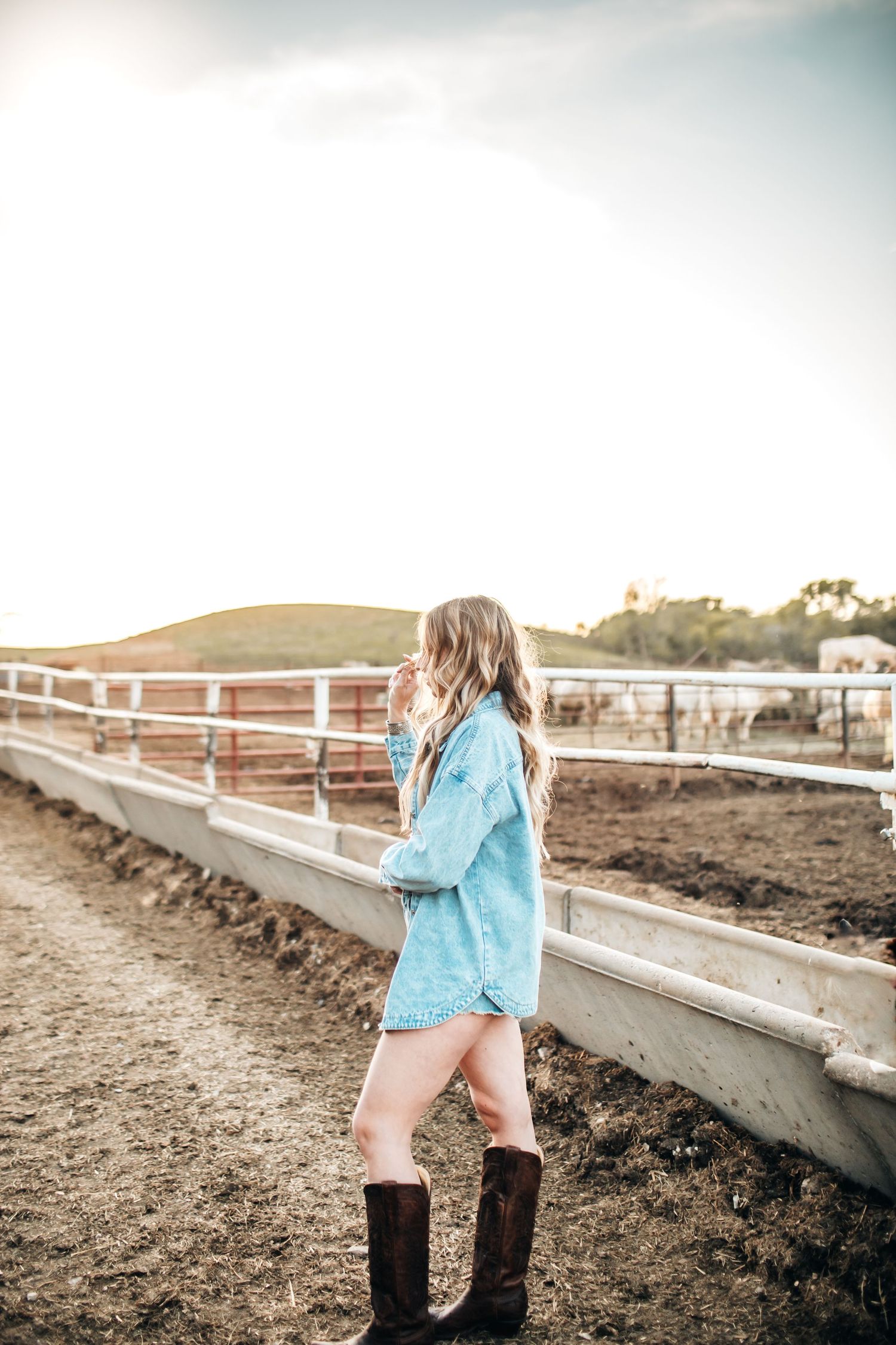 Woman in denim jacket and boots stands by white fence in outdoor photoshoot.