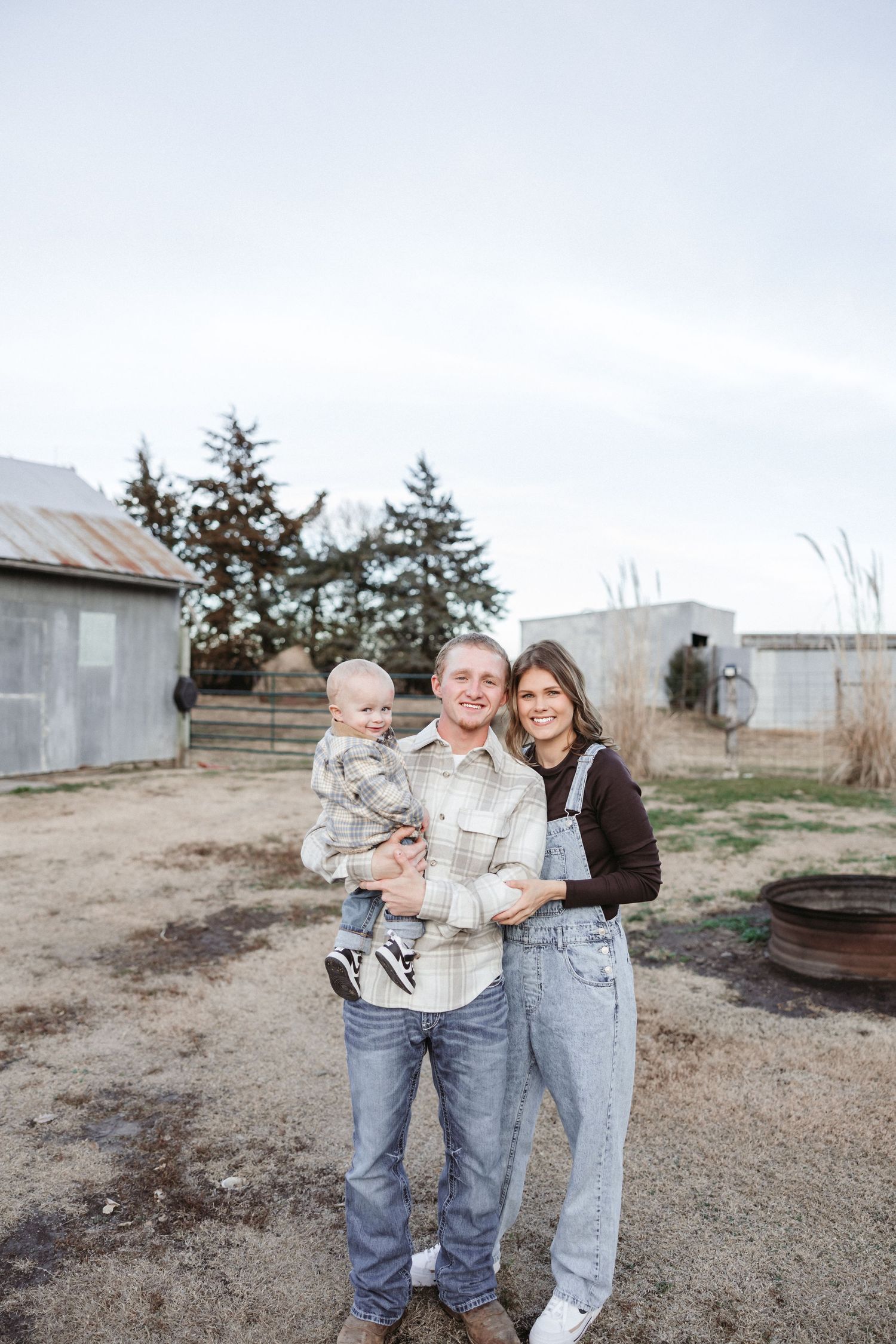A family poses together outdoors on a rural property with rustic buildings in the background.