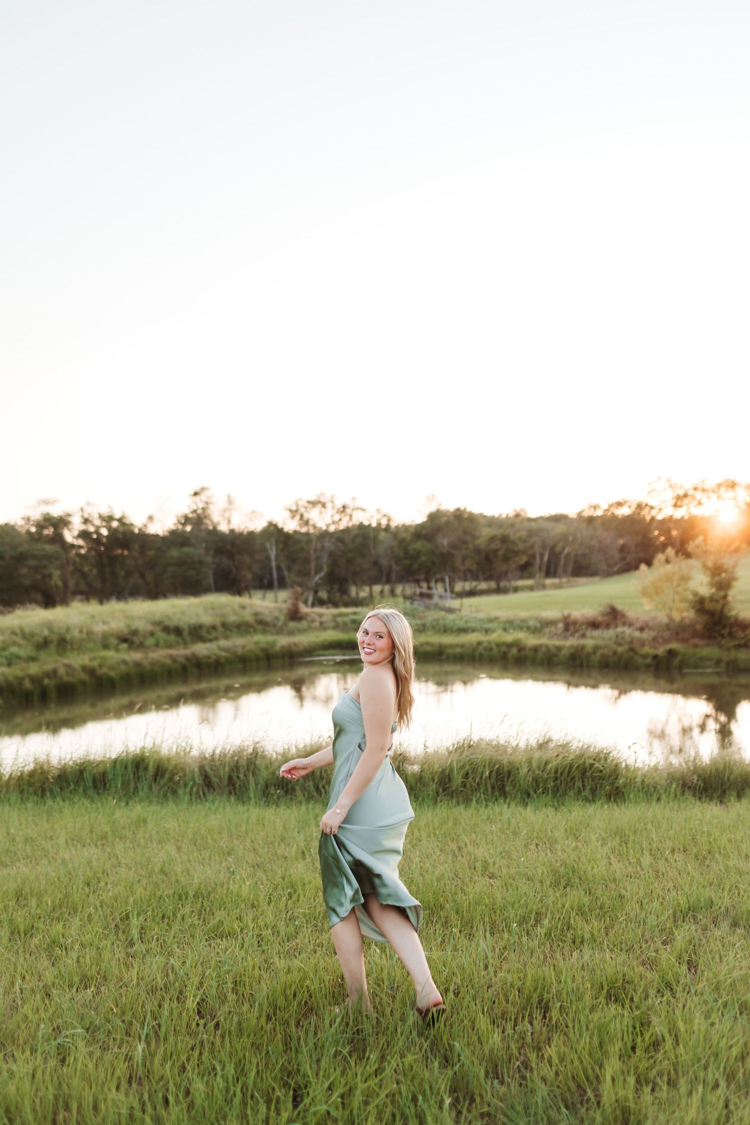 Woman in sage green dress walking barefoot in lush meadow near pond at golden sunset.
