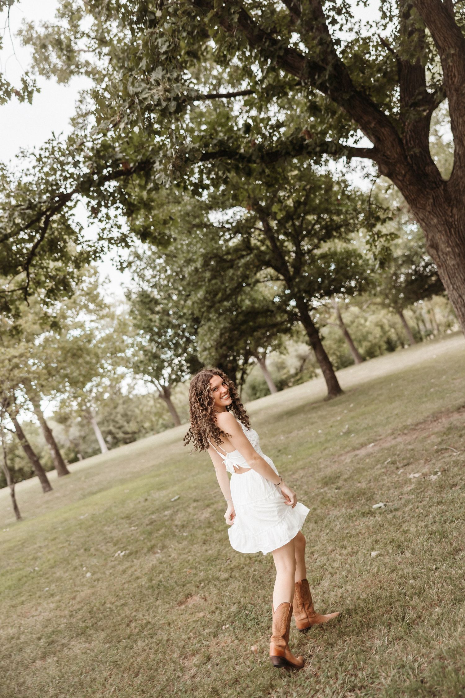 Young woman in white dress and cowboy boots twirling in a lush green park under large trees.