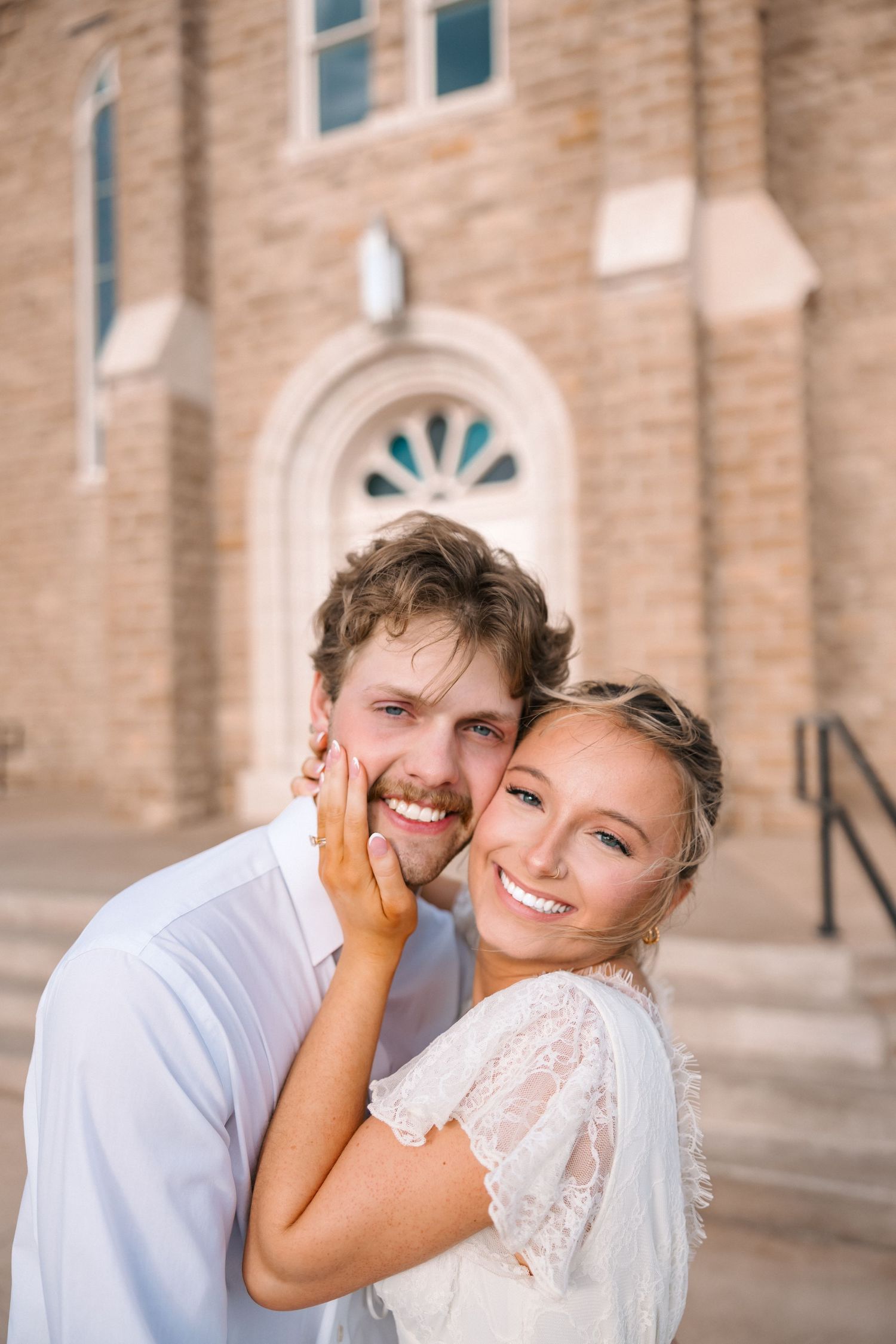 Smiling bride and groom pose together outside a stone church, wearing white wedding attire.
