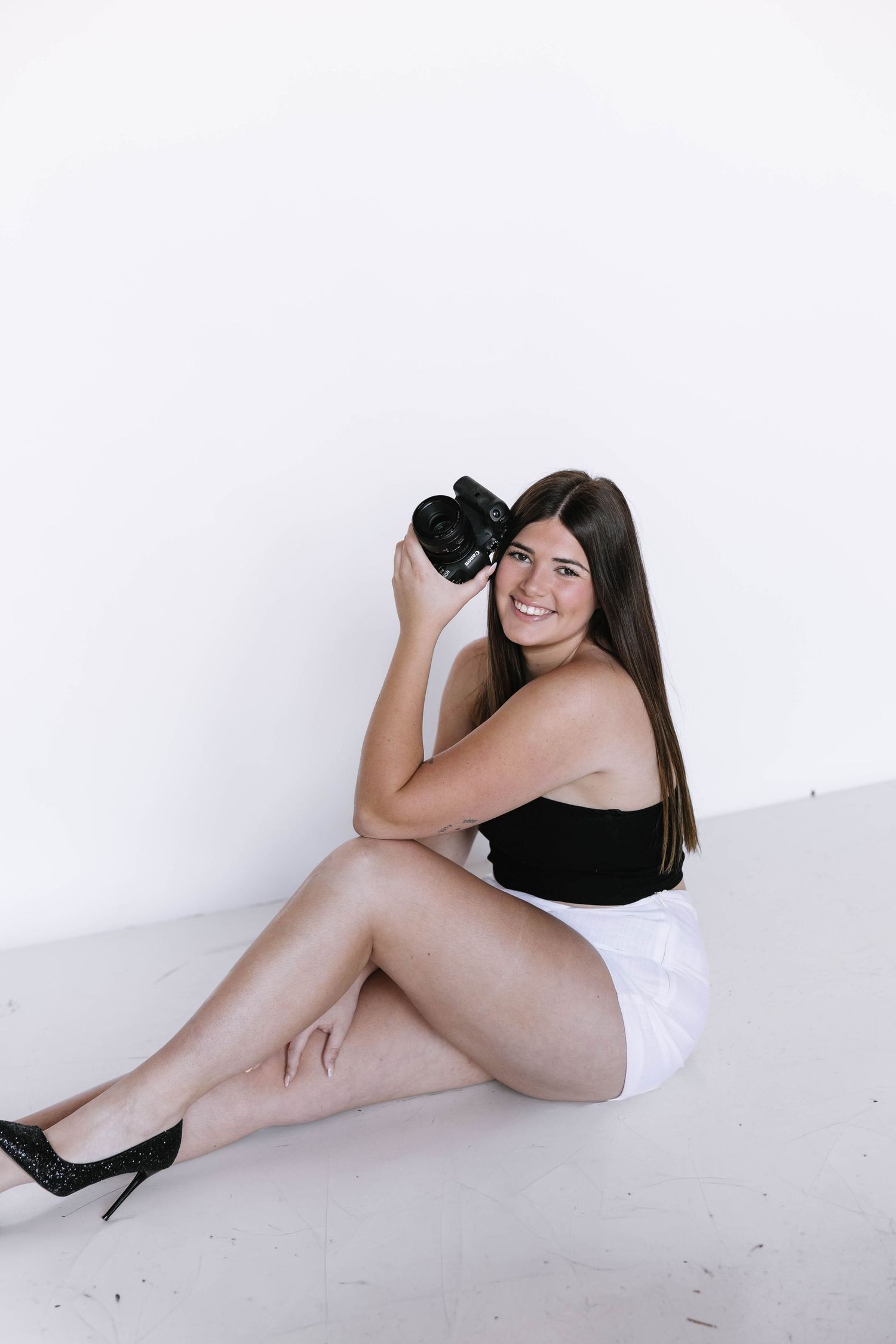Young woman sitting on floor holding a camera, wearing black top and white shorts, smiling at camera.