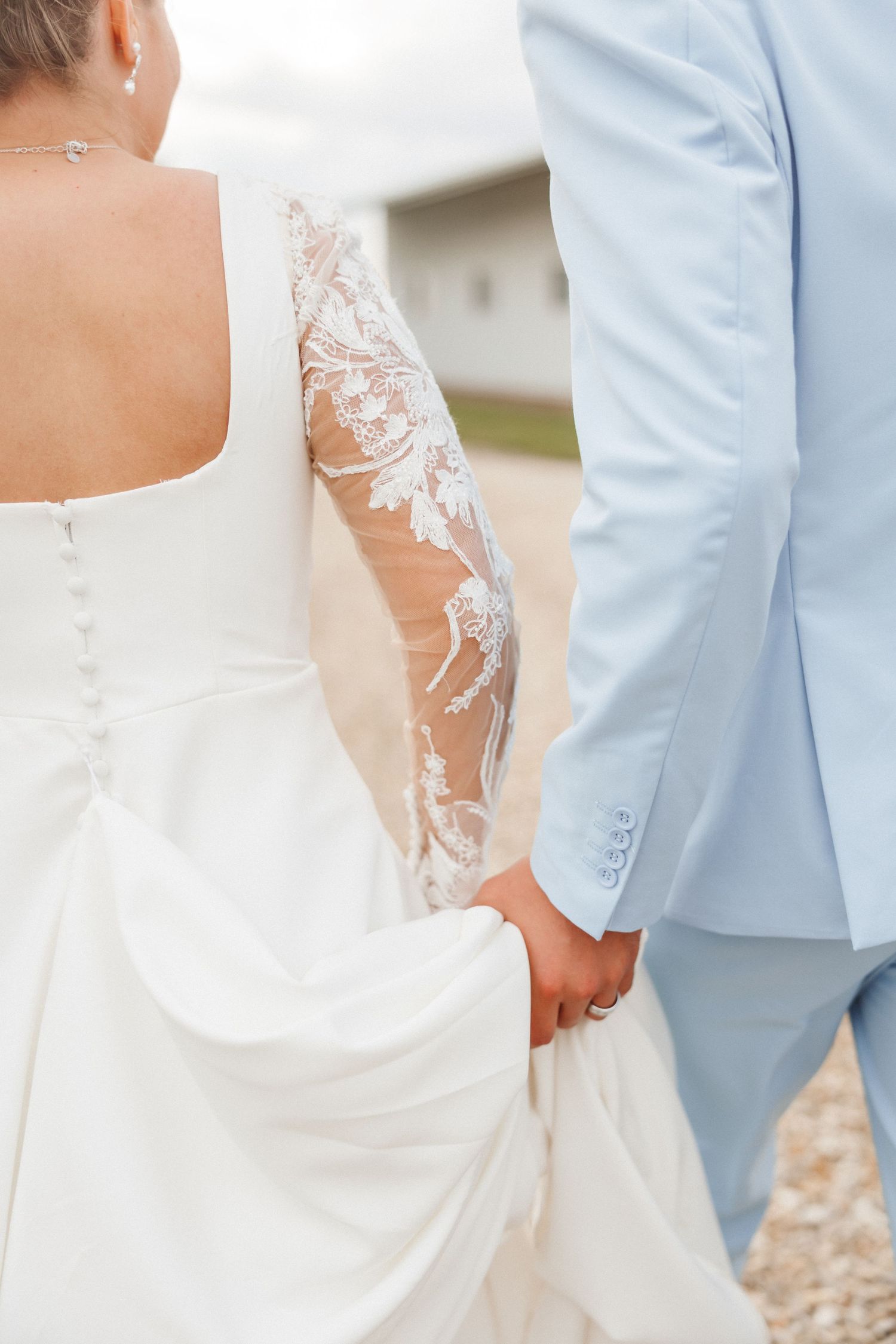 Bride and groom holding hands, showcasing lace wedding dress sleeve and light blue suit jacket.