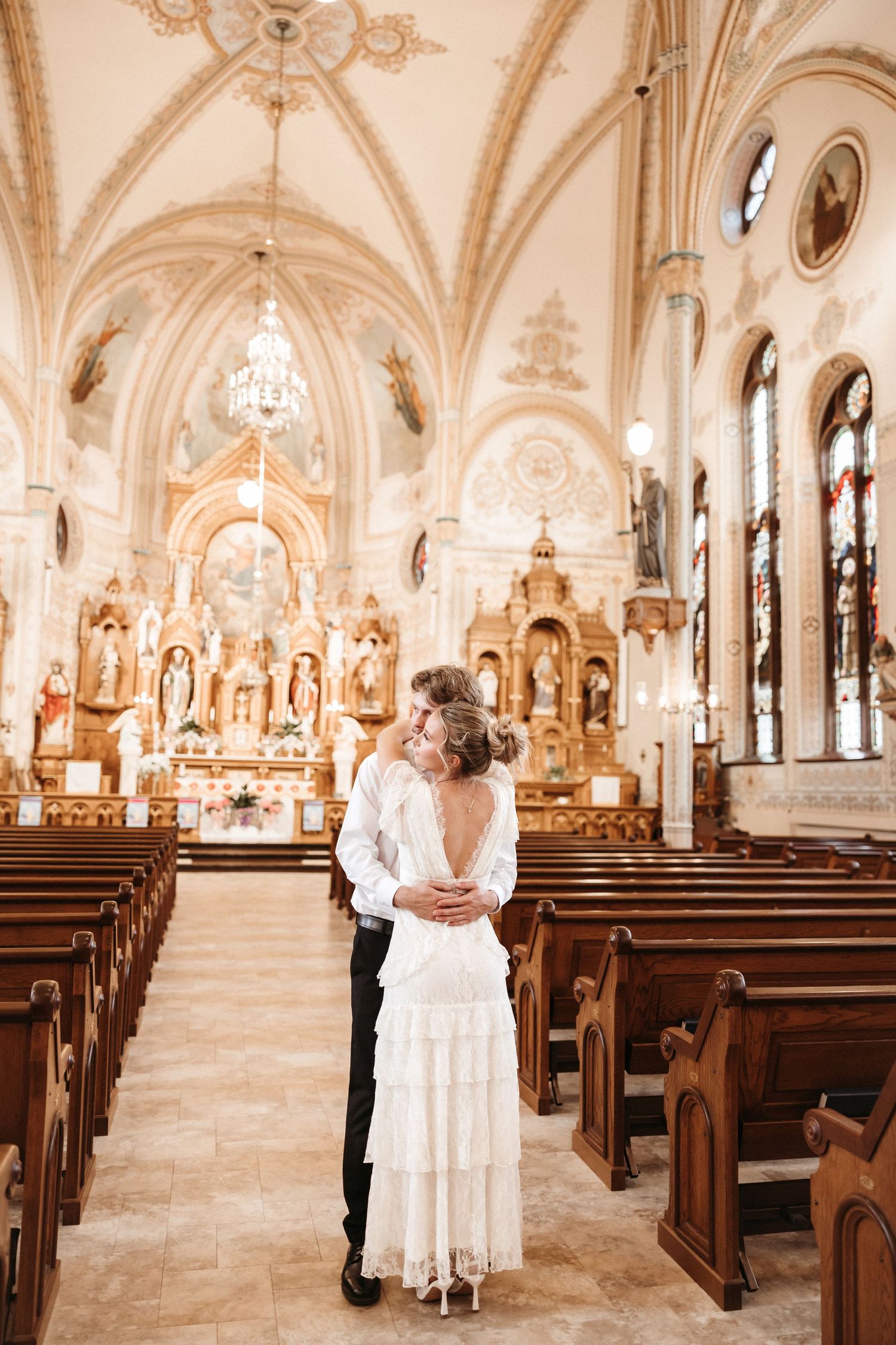 Bride and groom embracing in an ornate Catholic church with golden altar, vaulted ceilings, and wooden pews.