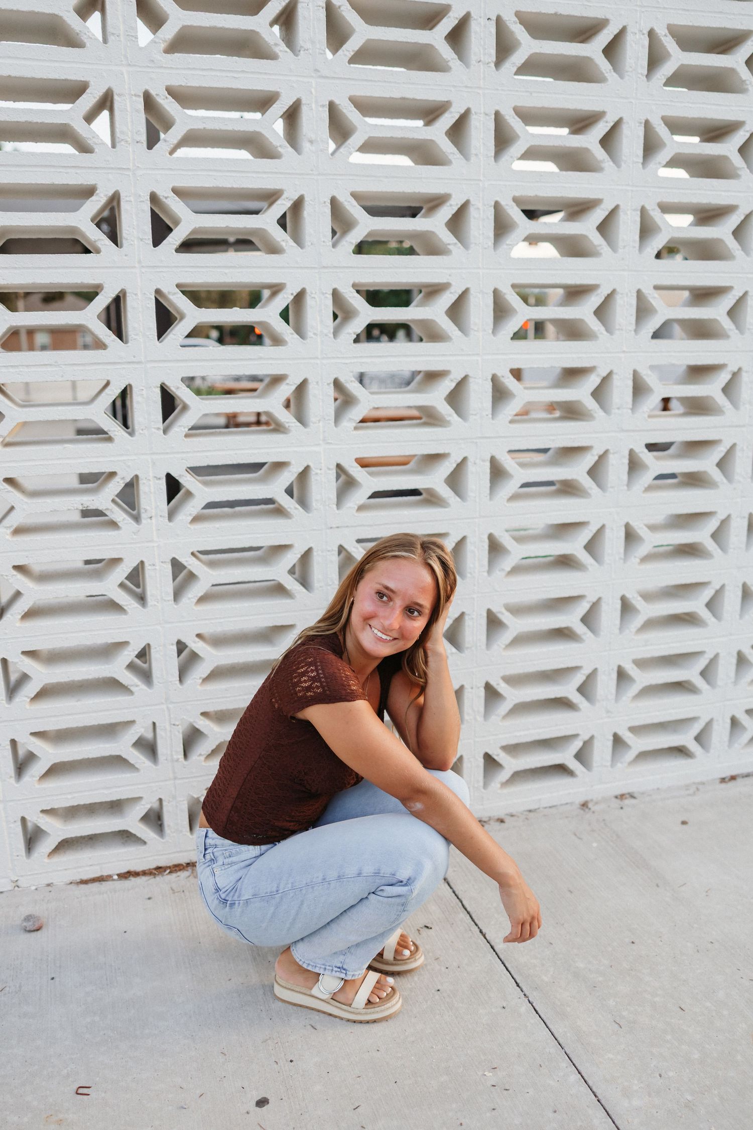 Young woman crouching in front of a white decorative lattice wall, wearing a brown top and light blue jeans.