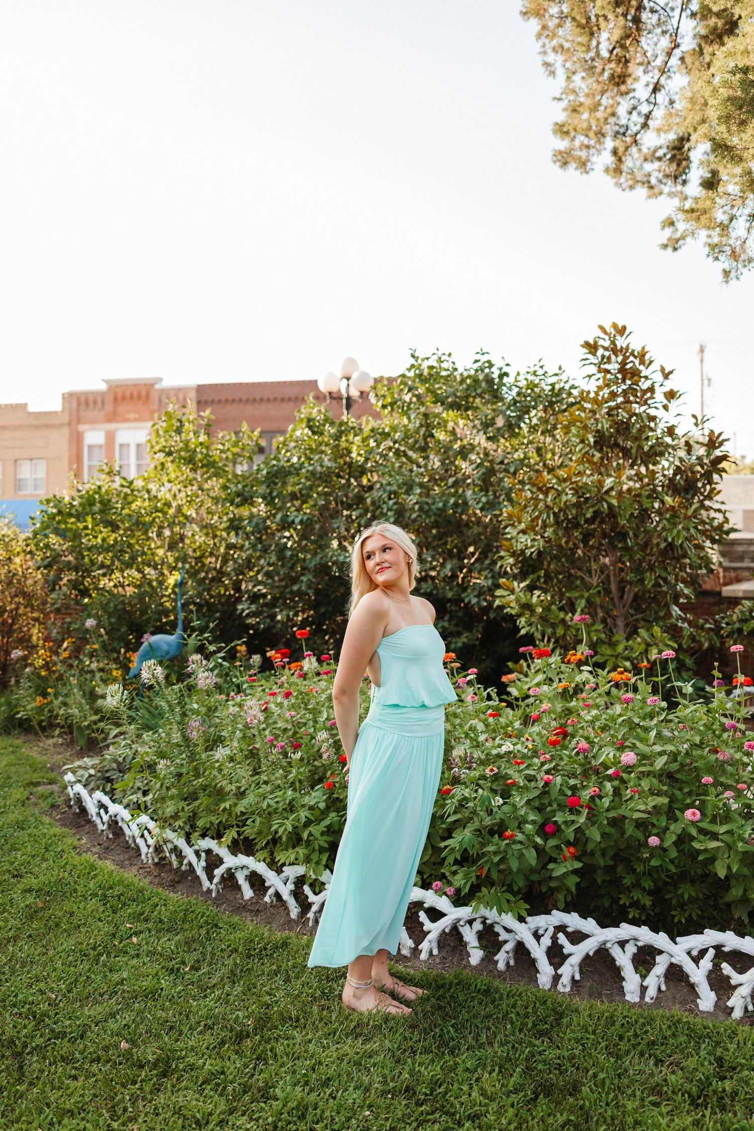 Woman in mint green strapless maxi dress standing barefoot in a colorful garden with red flowers and lush greenery.