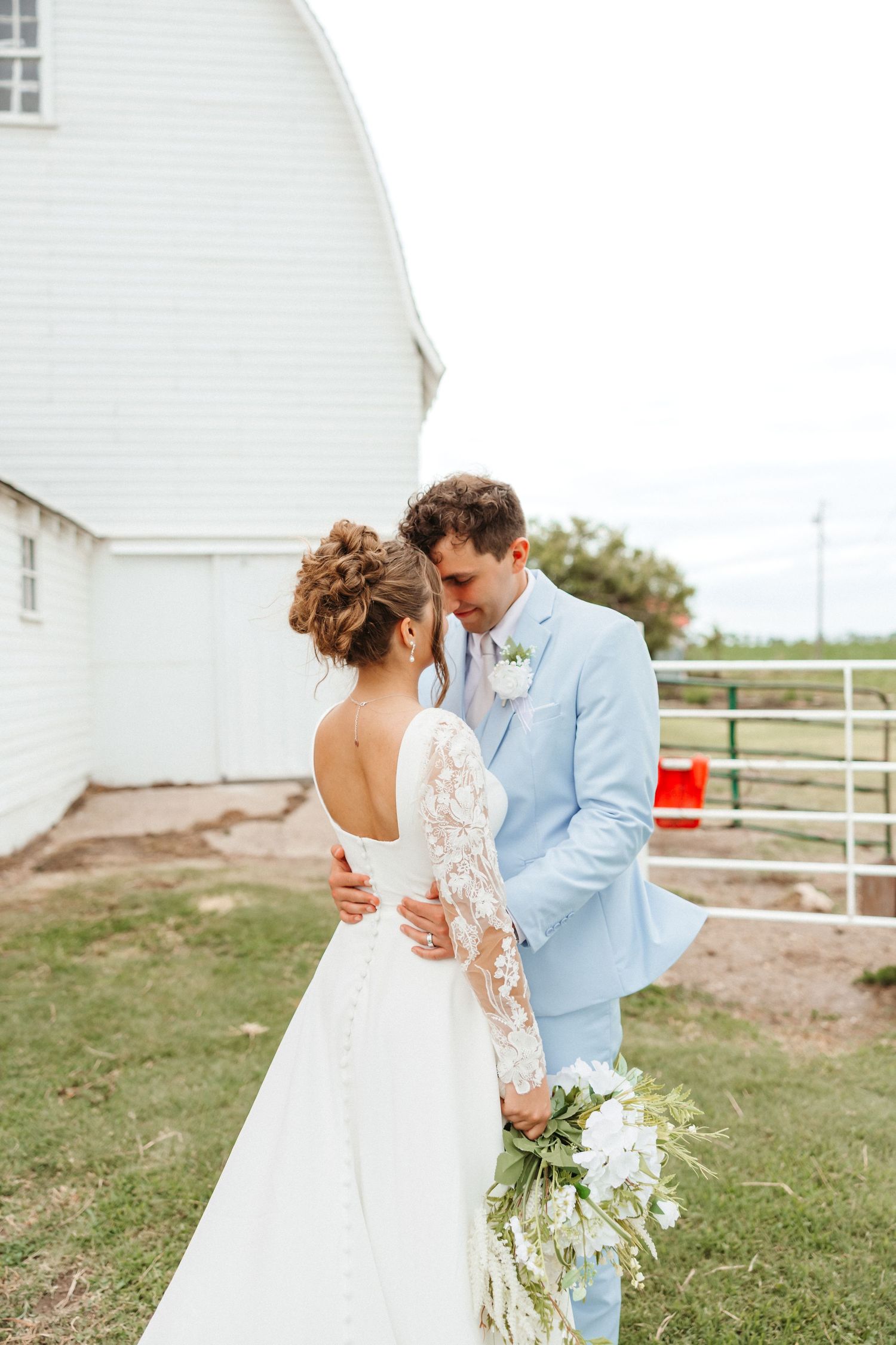 A bride and groom share a tender moment outside a white barn, she in lace gown, he in light blue suit holding bouquet.