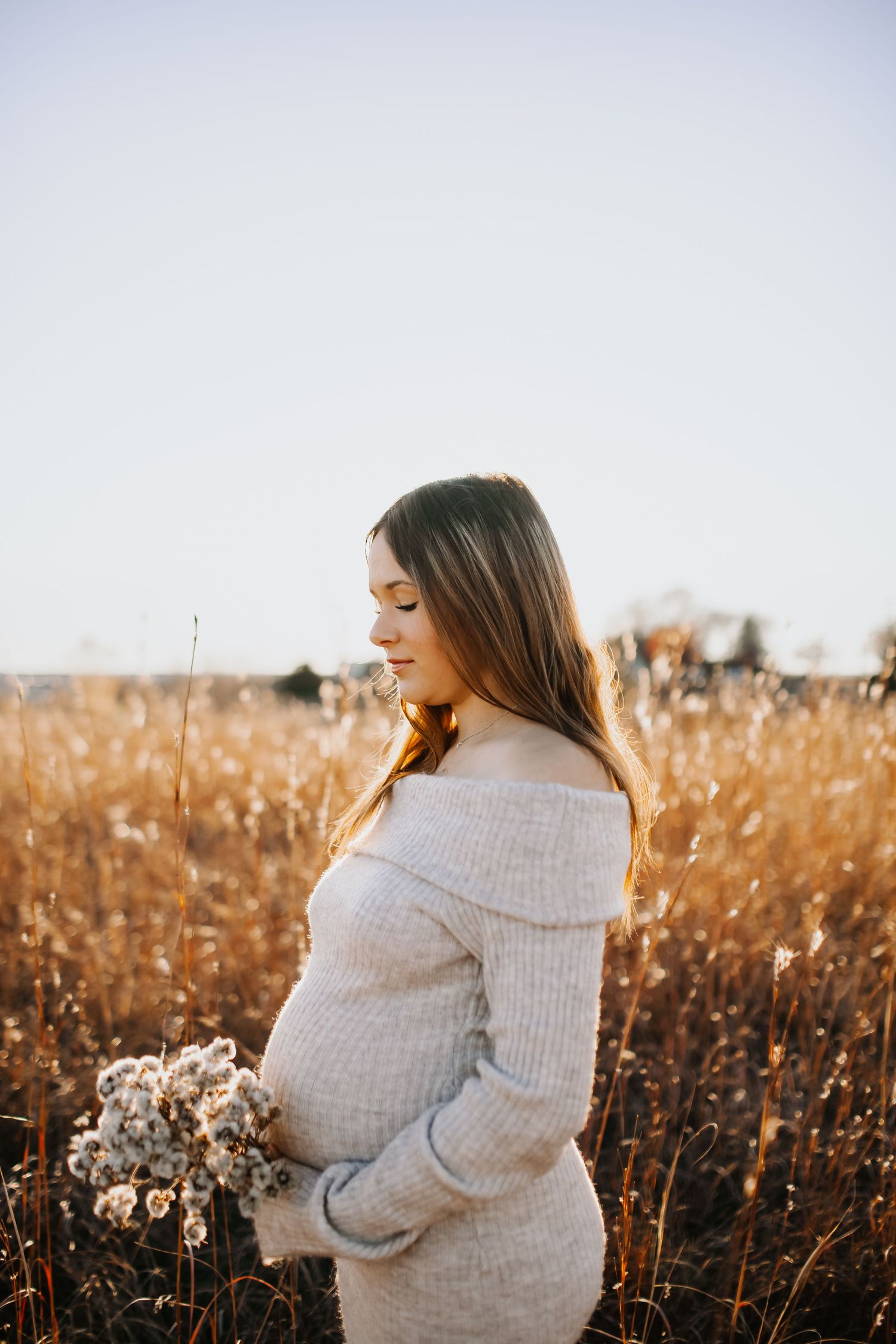 Expectant mother in cozy sweater dress holding dried flowers in golden autumn field at sunset.