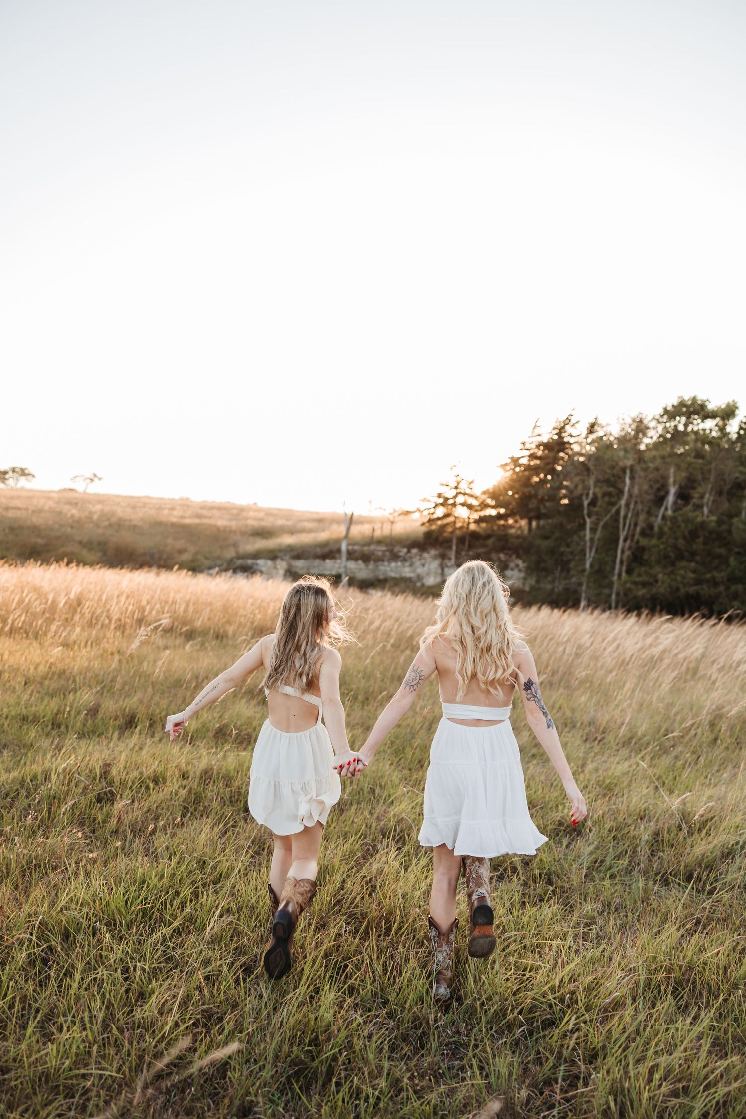 Two women in white dresses and cowboy boots hold hands walking through a golden grass field at sunset.
