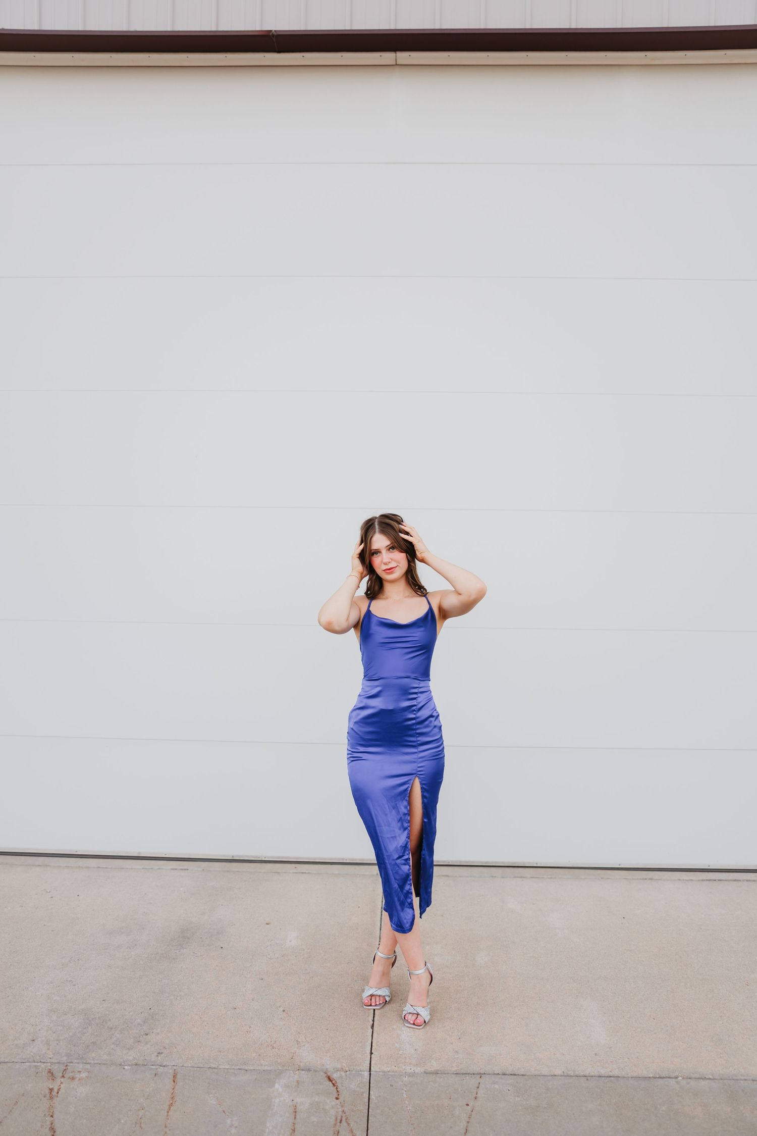 Woman in blue satin midi dress with slit and heels posing in front of white garage door.