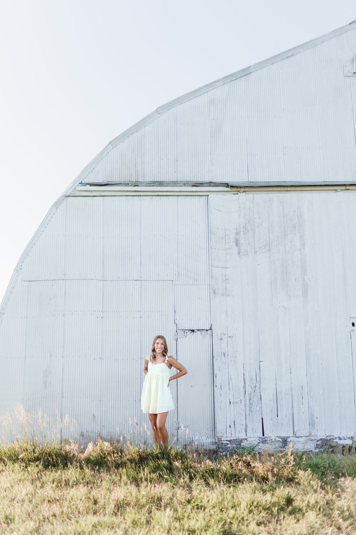Young woman in white sundress standing before a large white curved barn in a grassy field.