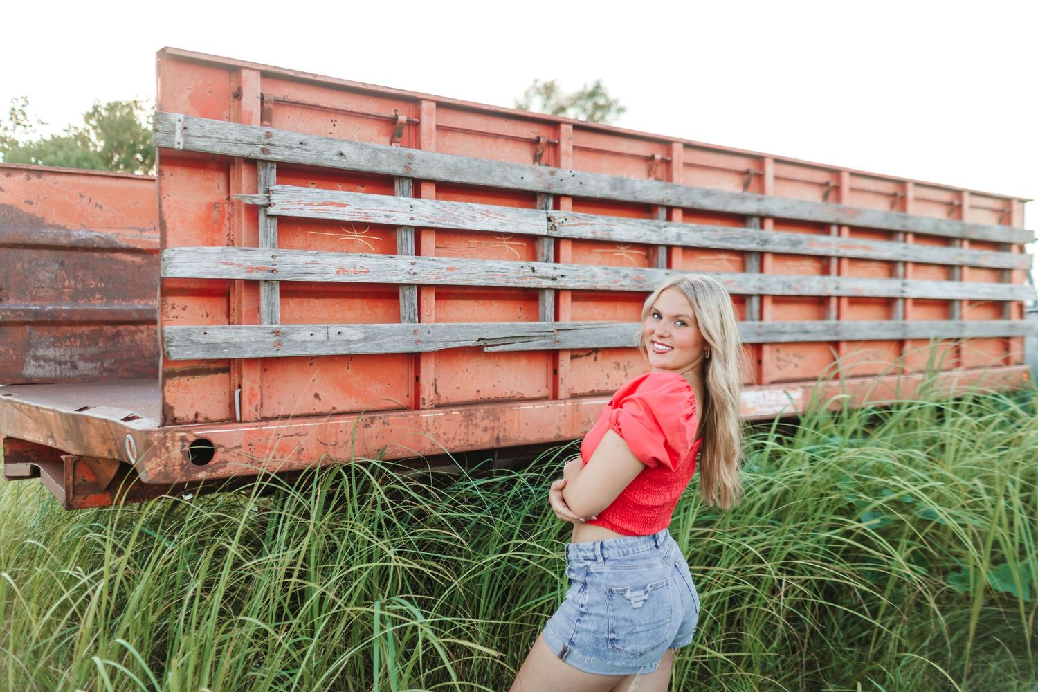 Woman in coral top and denim shorts poses playfully against orange farm equipment.