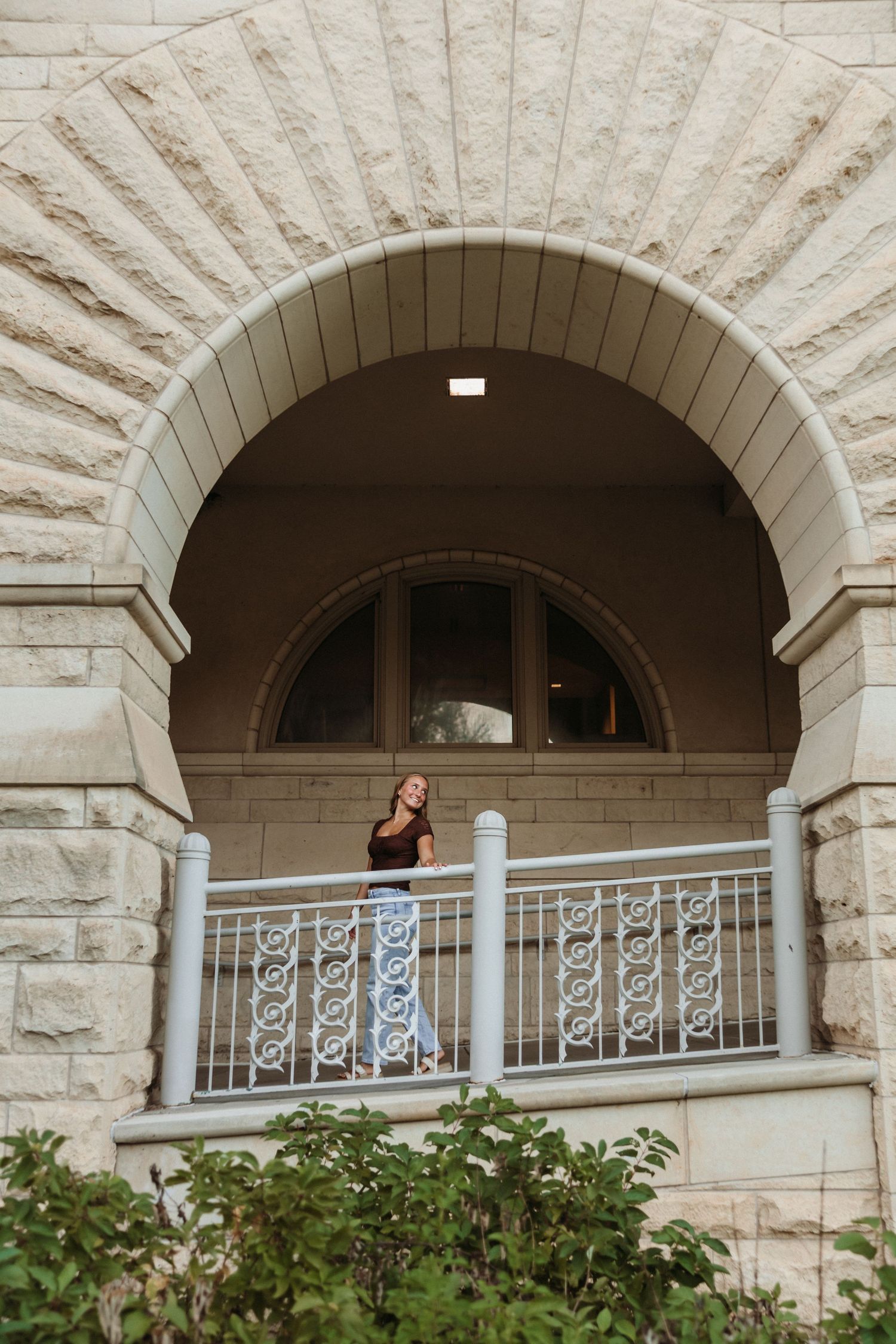 Woman standing on ornate stone arch balcony with decorative iron railing at a historic building.