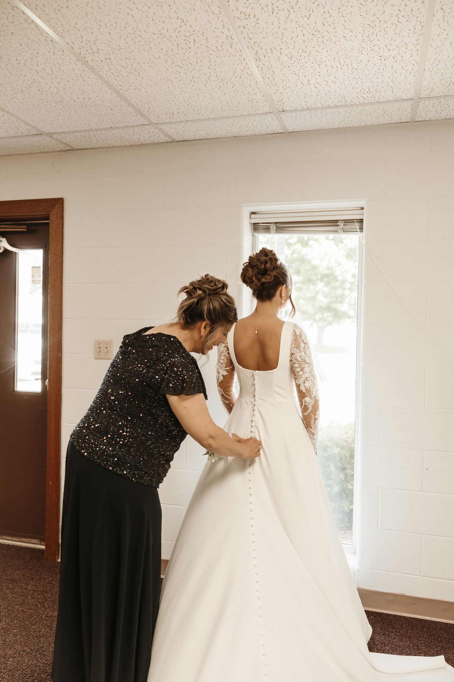 A woman in a black dress helps a bride in a white gown with her dress buttons before the wedding ceremony.