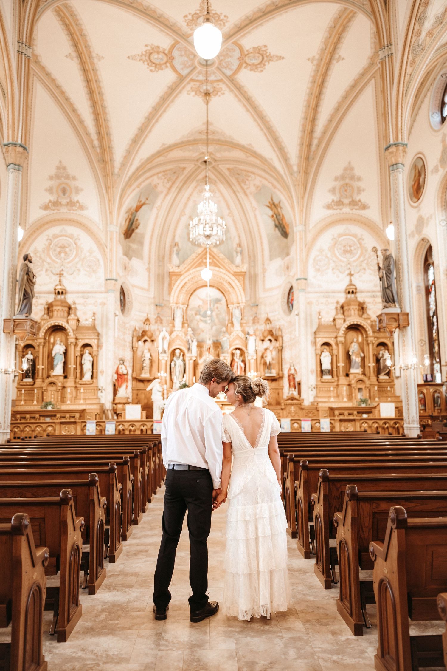 Bride and groom standing hand in hand at the altar of an ornate white Catholic church with vaulted ceilings.