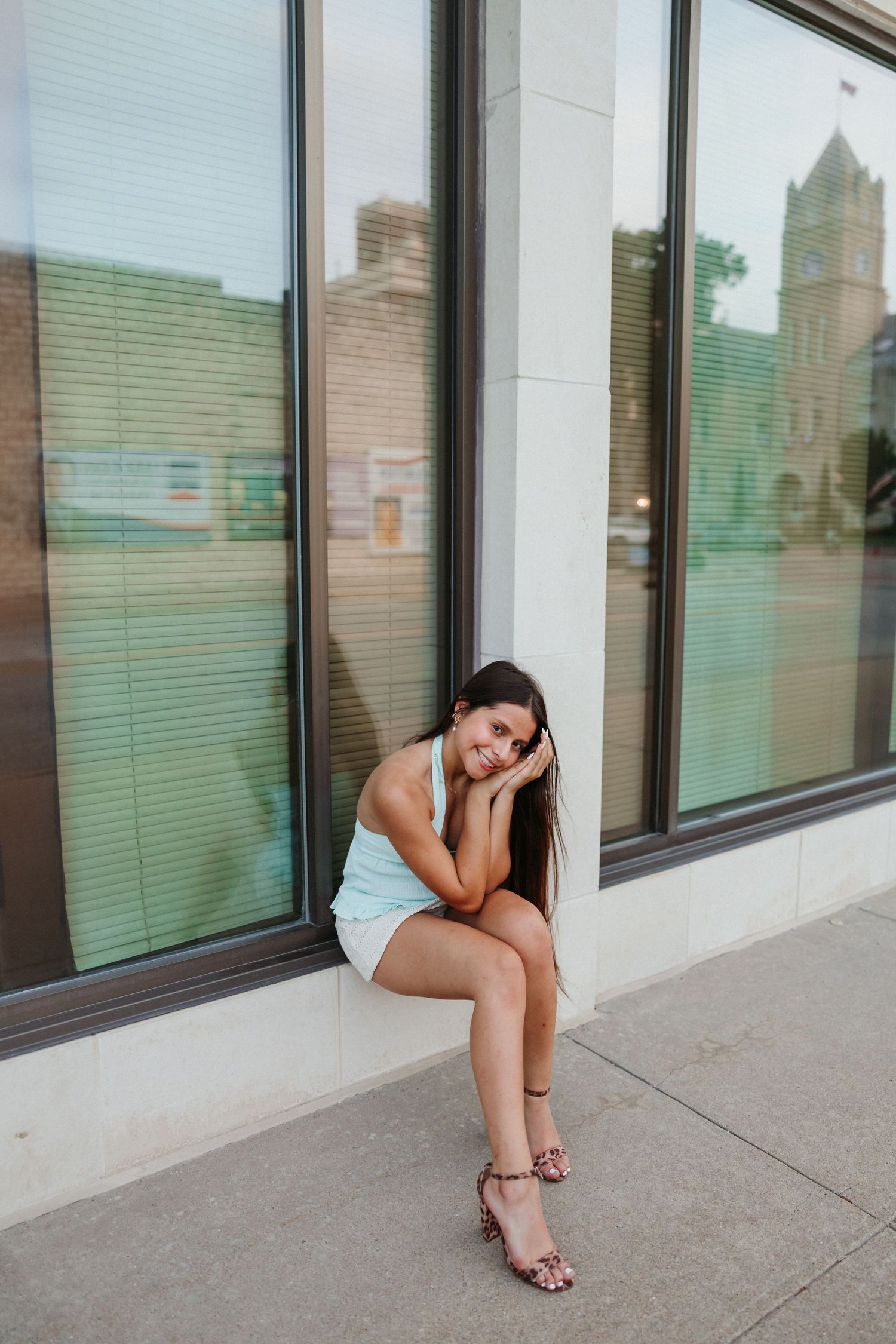 Young woman in white dress and leopard heels sits against a building window on a sunny urban sidewalk.