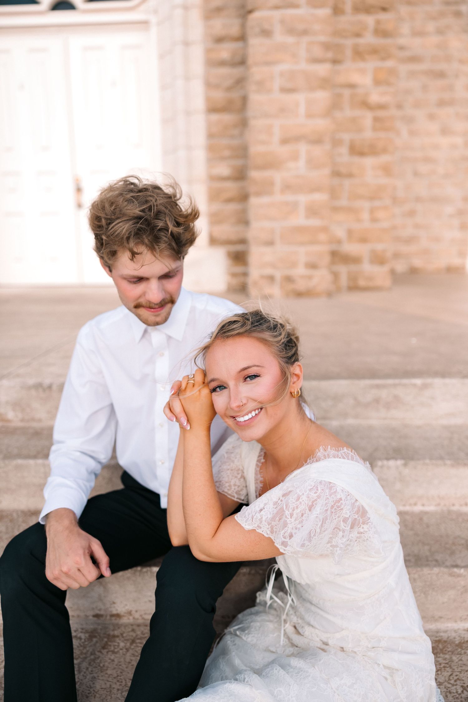 A smiling bride in lace dress sits on steps with groom in white shirt, posing together outside a stone building.