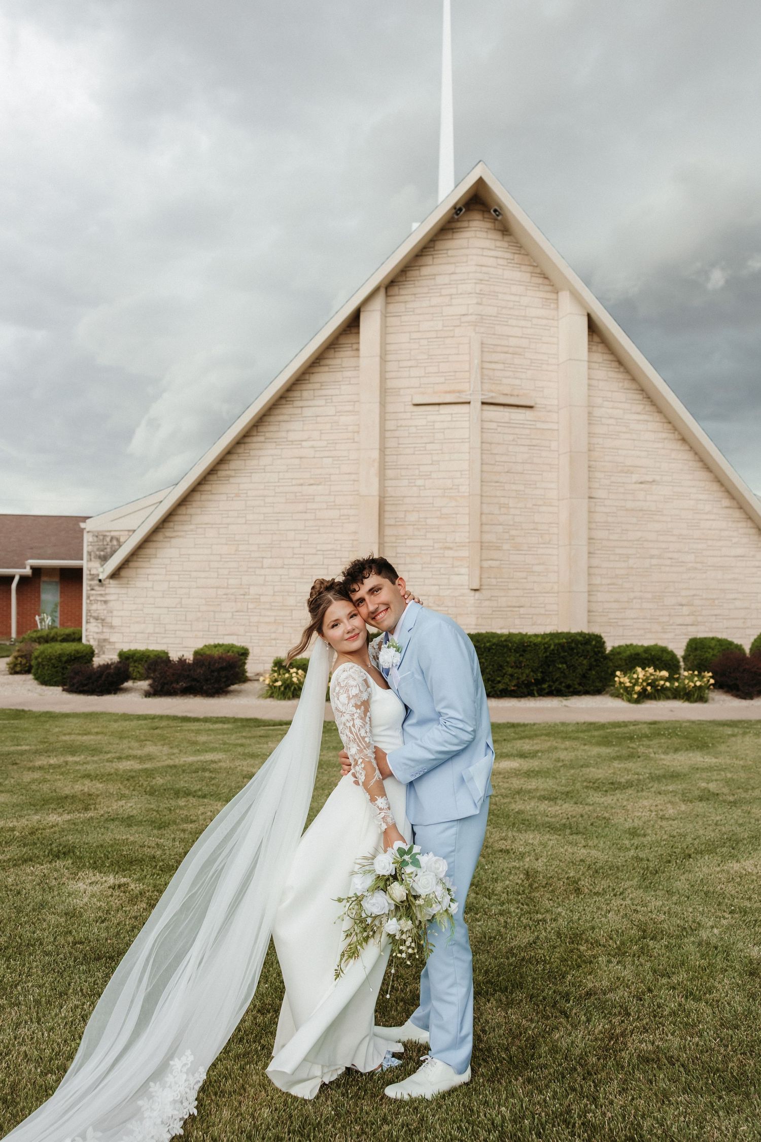 Bride and groom pose in front of a brick church with a cross, bride holding white bouquet, veil flowing in the wind.