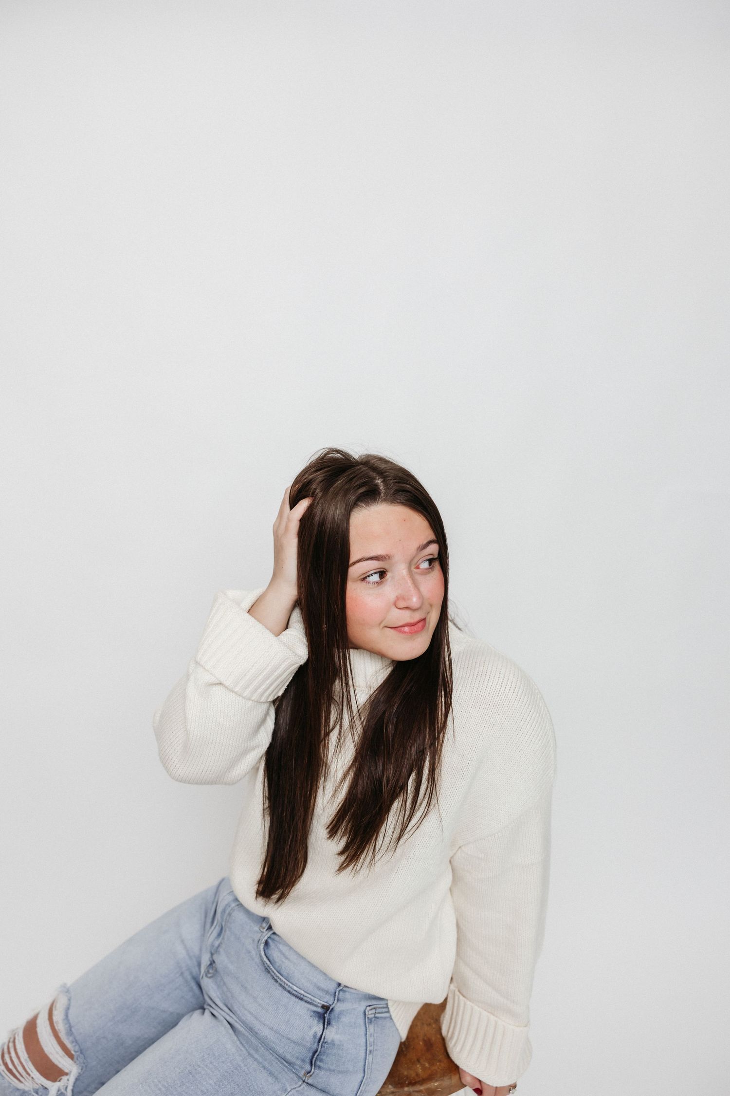 A casual portrait against white background wearing cream sweater and distressed jeans while touching hair.