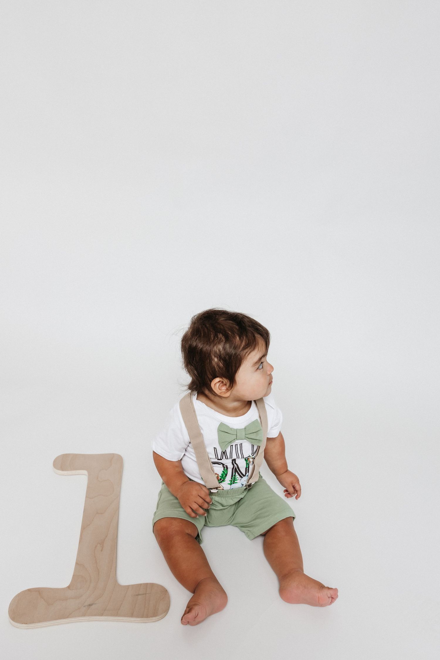 Baby in green and white outfit near wooden number one on white background.