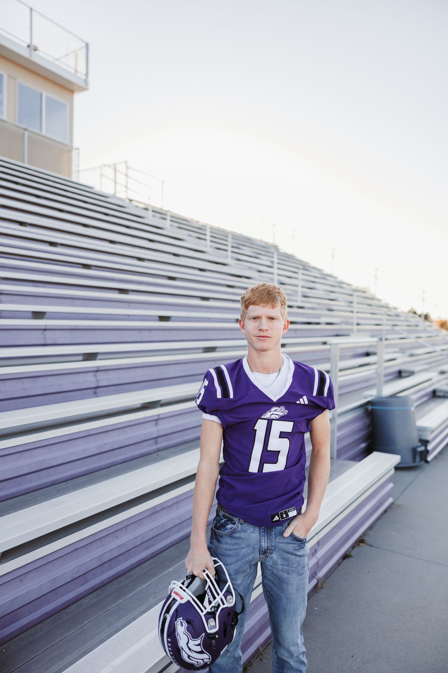 High school athlete in purple football jersey poses on stadium bleachers.