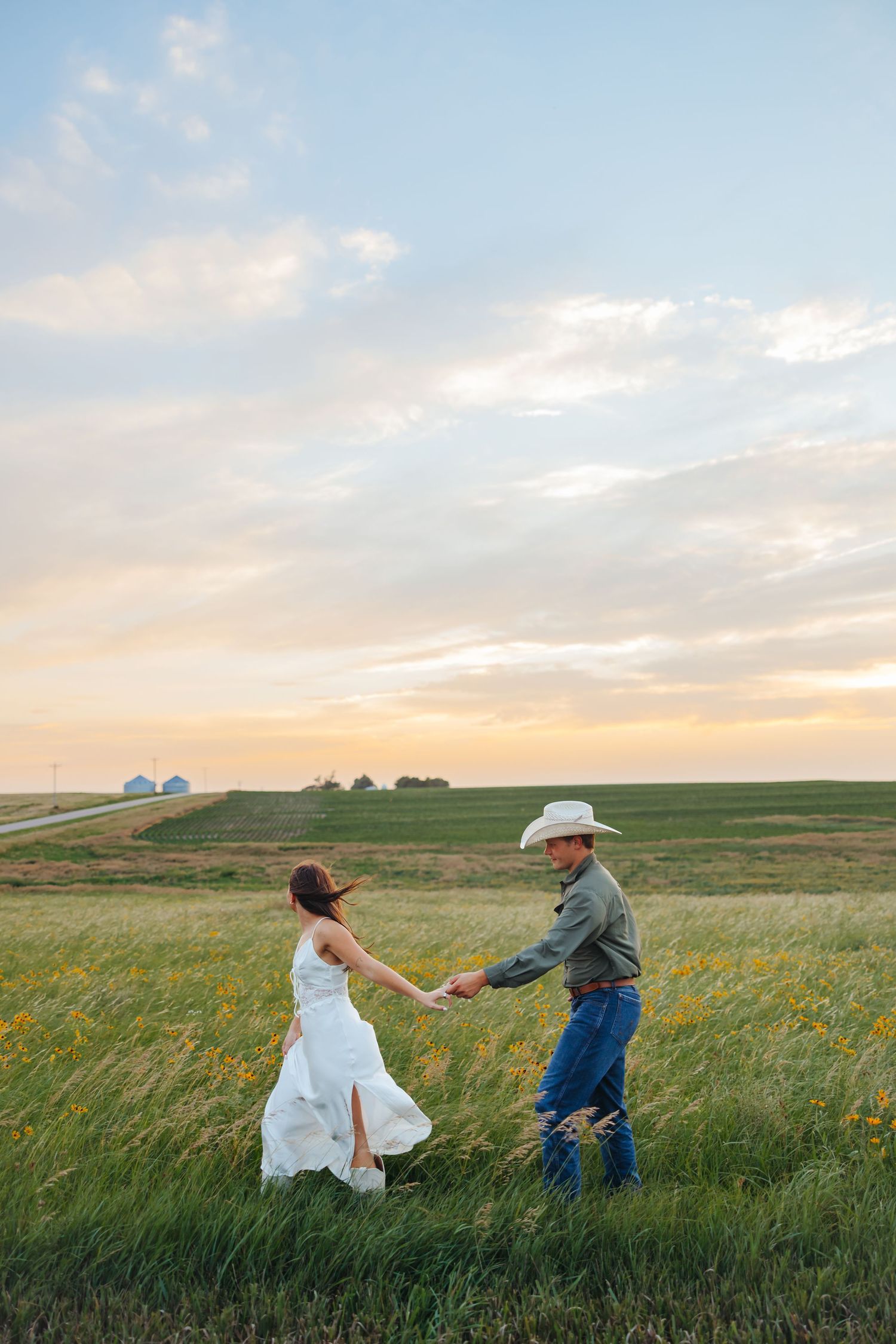 Couple in wedding attire holding hands in a green field at sunset, groom in cowboy hat and bride in white dress.