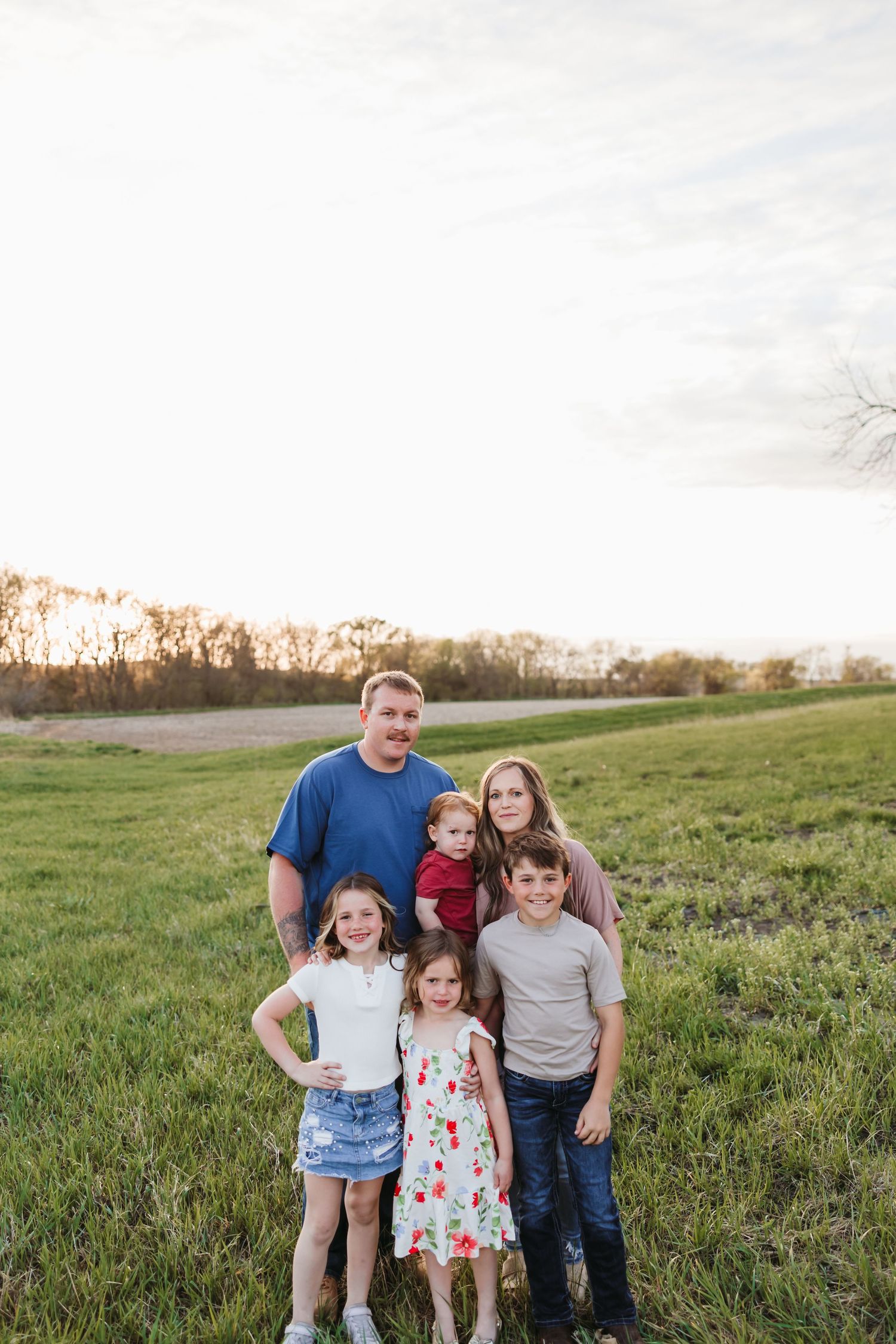 Family of five posing in a green field at golden hour, parents with three children smiling outdoors.