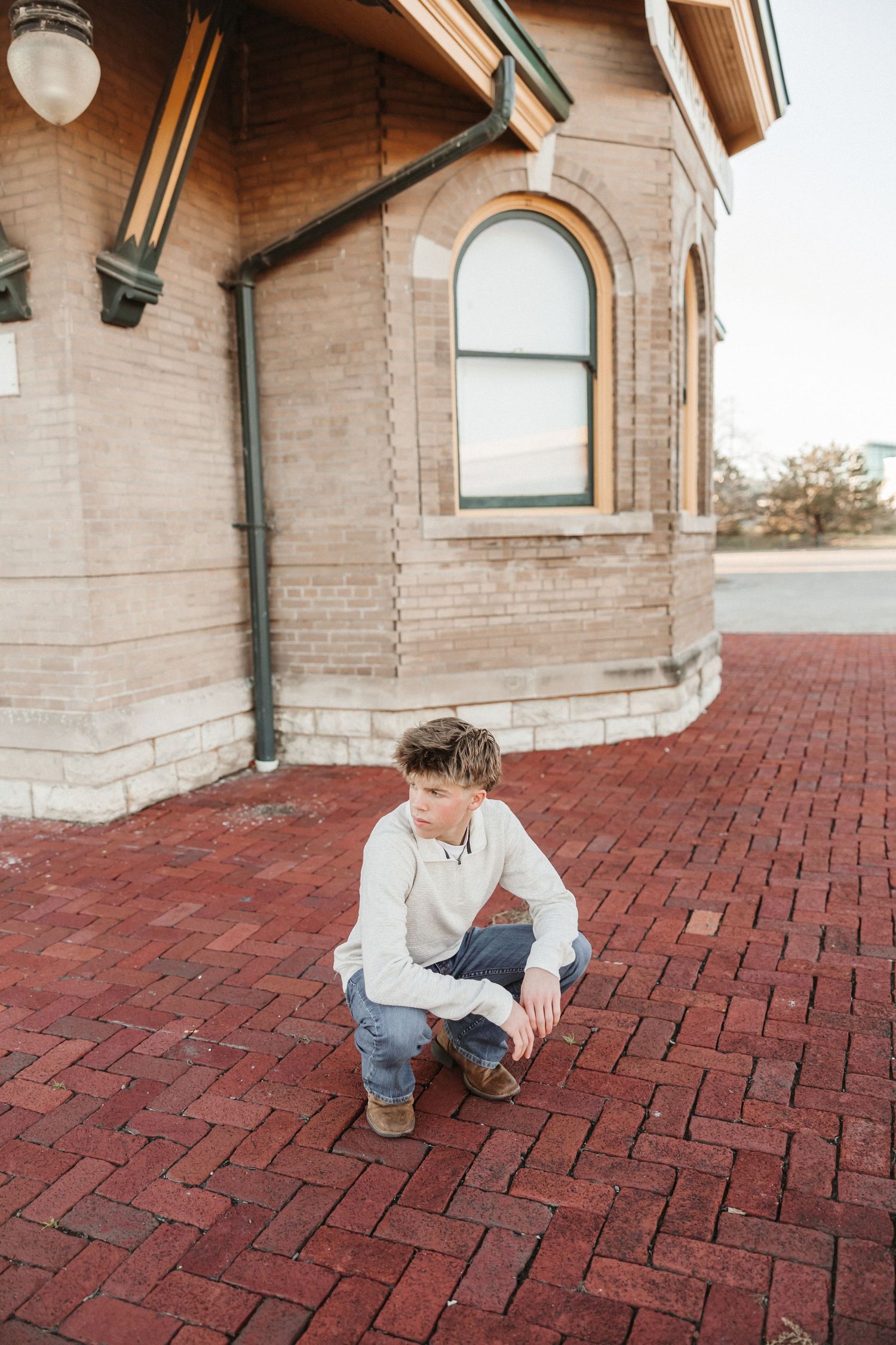 Person crouches on red brick pavement near stone building.