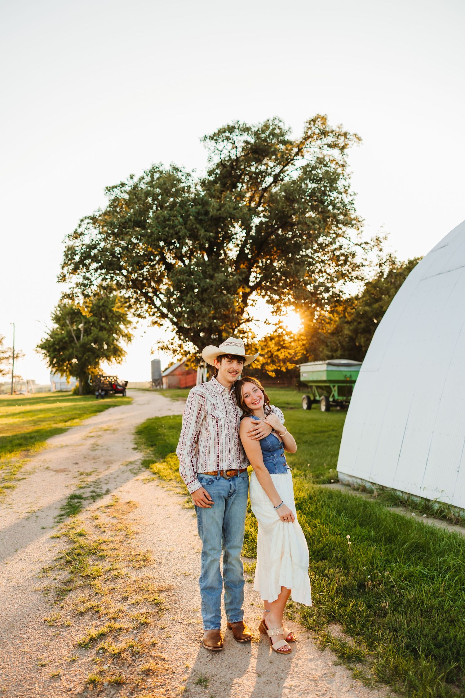 Couple in cowboy hats embracing at golden hour on a farm, with a gravel path, barn, and large tree in the background.