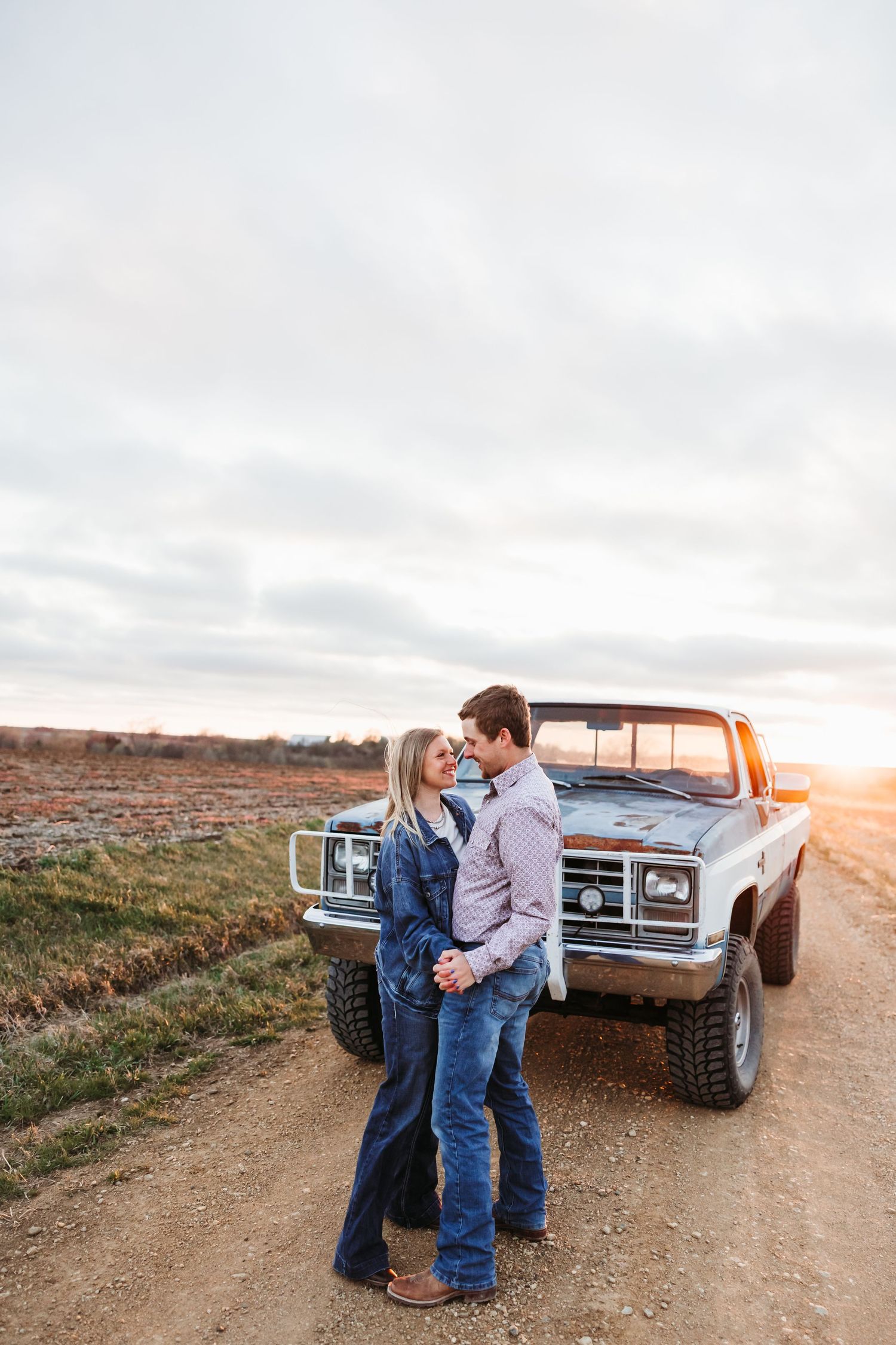 Couple embracing near a vintage pickup truck on a dirt road at sunset in a rural field.