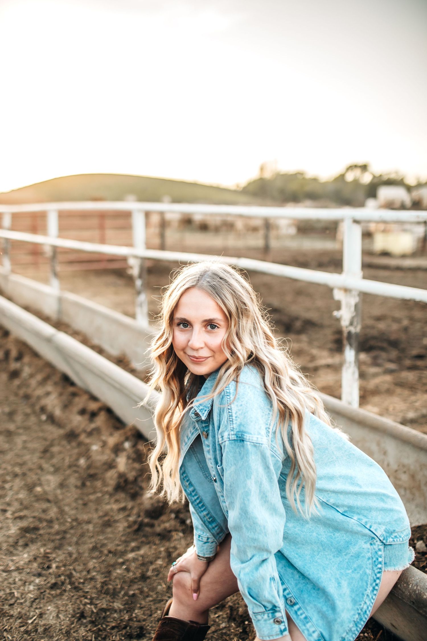 Woman outdoor portrait by white fence featuring light denim jacket and flowing hair.