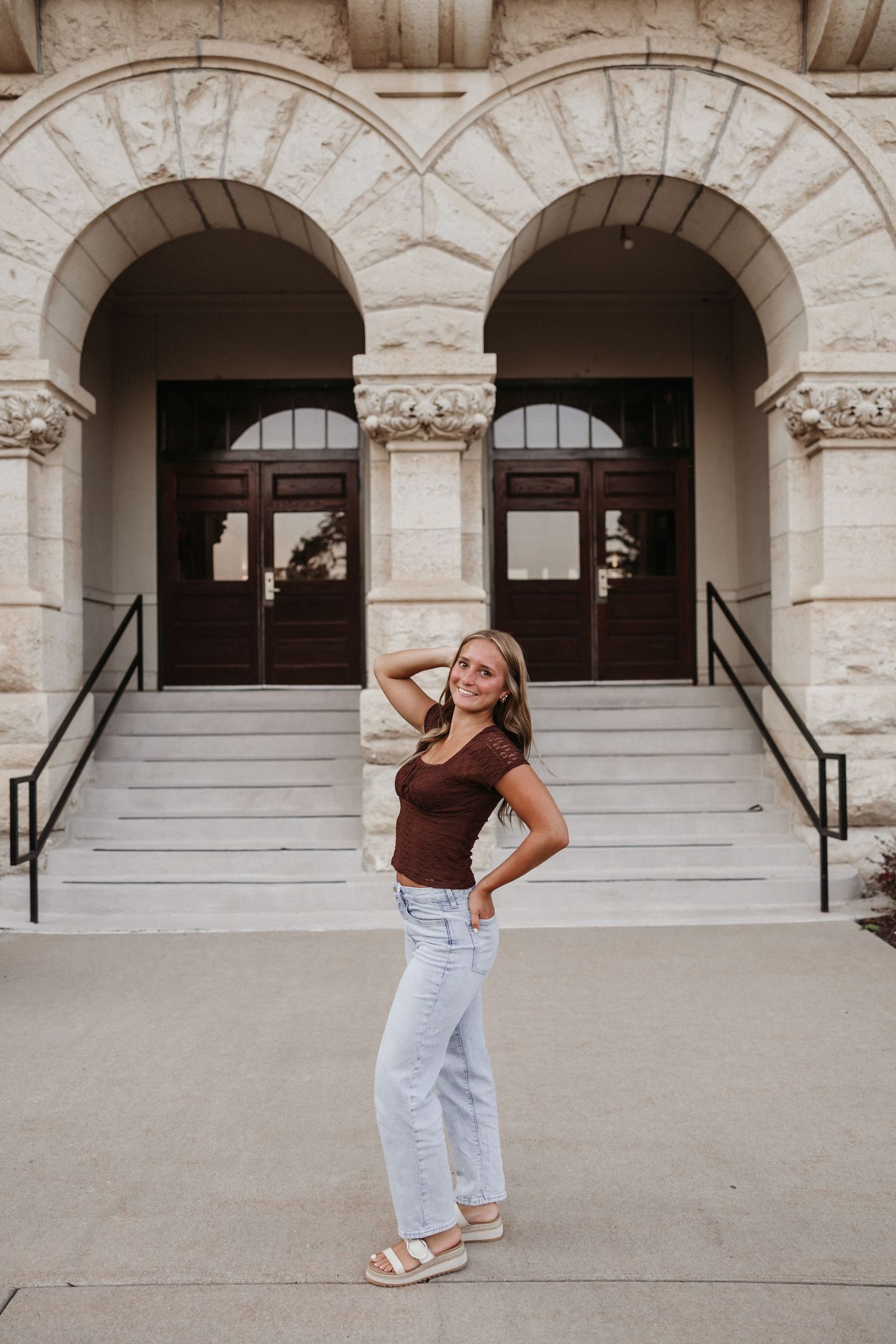 Young woman in brown top and light jeans poses smiling before grand arched stone building entrance with steps.