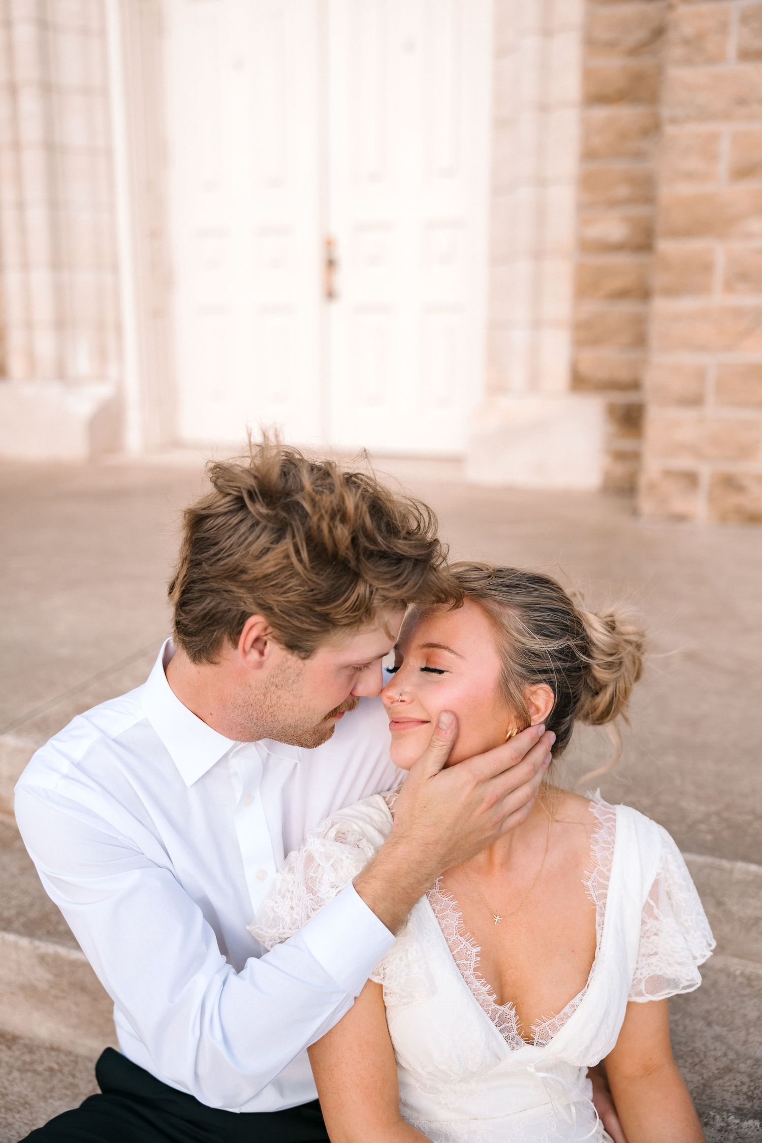 Groom tenderly holds bride's face while leaning in for a kiss outside a stone church.