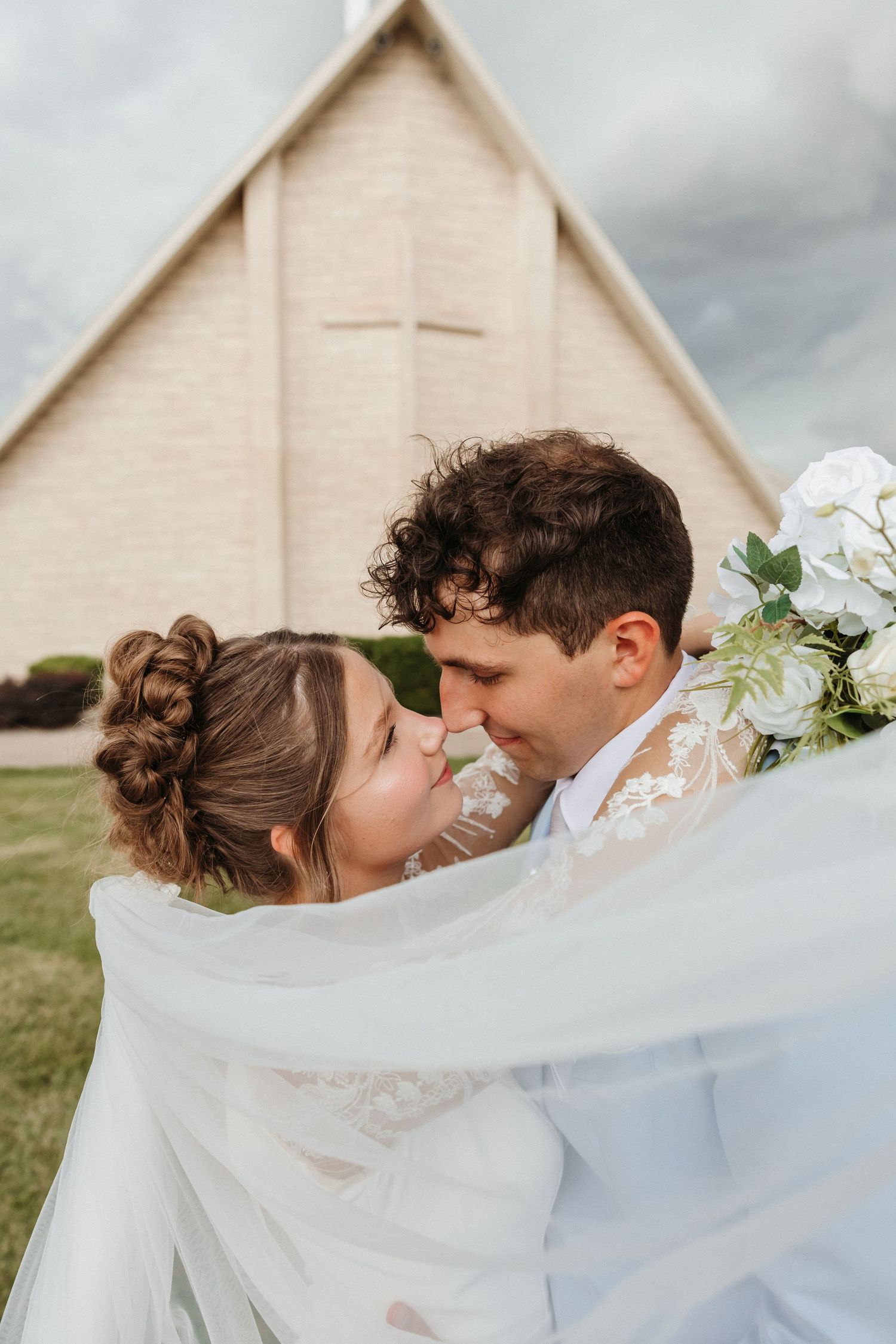 A bride and groom share a romantic moment outside a church, with her veil flowing and white bouquet in hand.