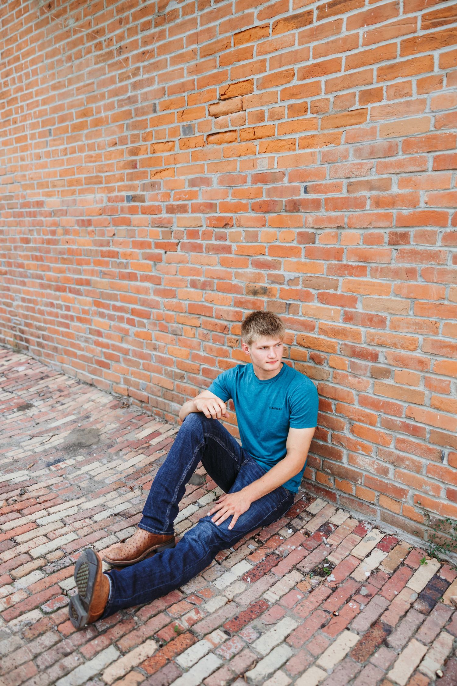 Young man in teal shirt and jeans sitting against a red brick wall on a brick paved floor.