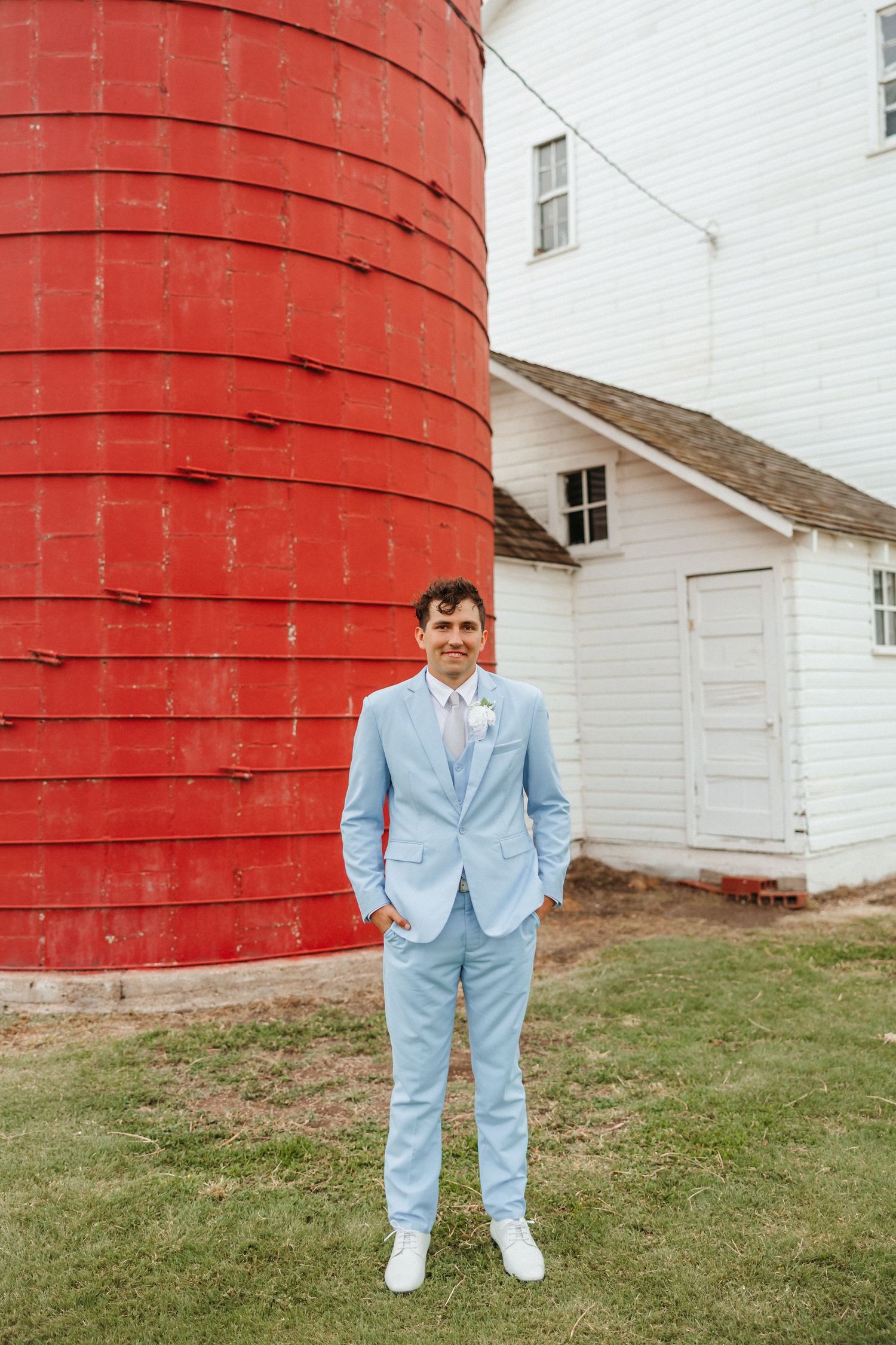 Groom in a light blue suit stands confidently in front of a large red silo and white barn building.