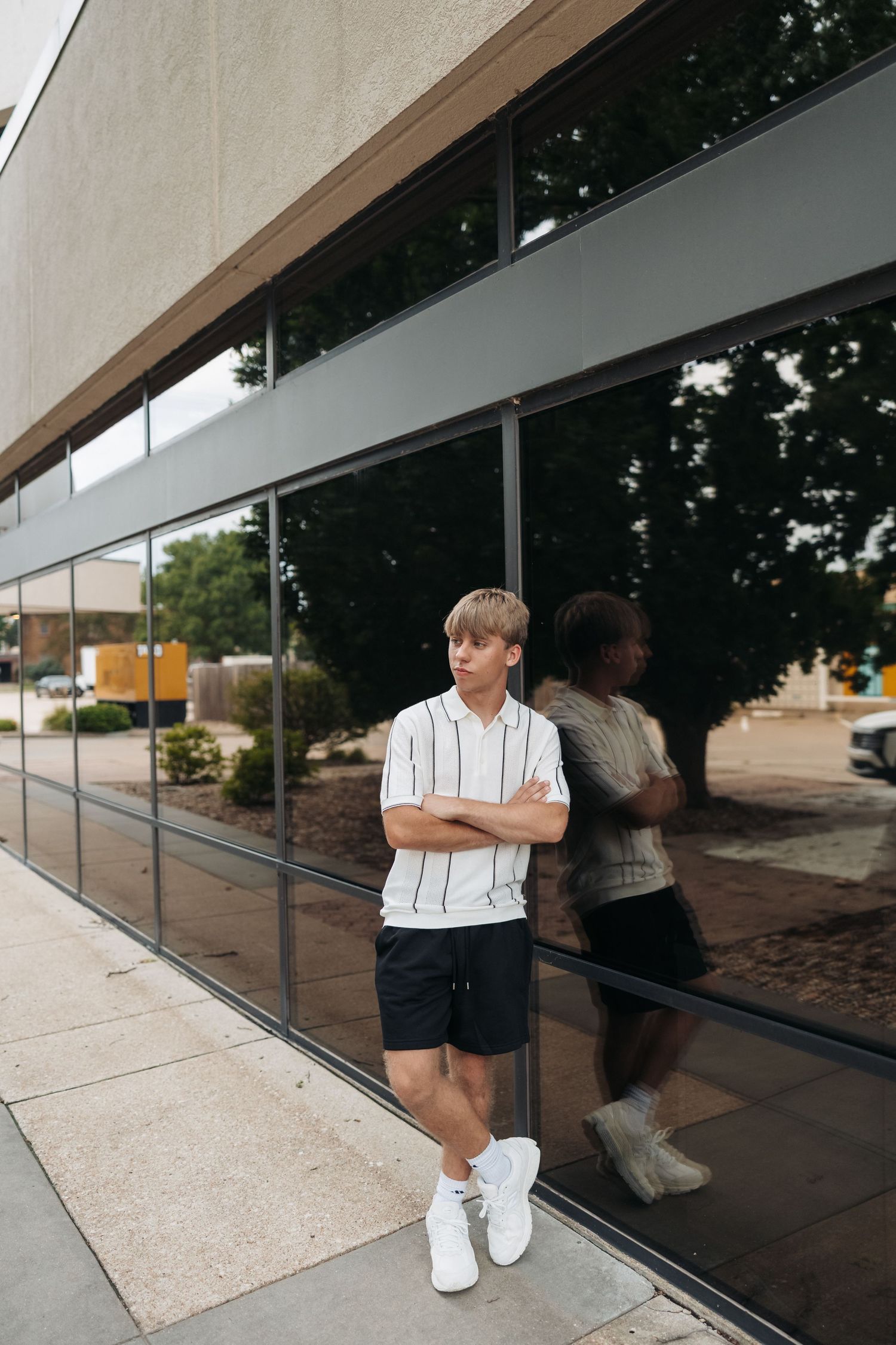 Young man in white striped polo and black shorts stands arms crossed against a modern glass building exterior.