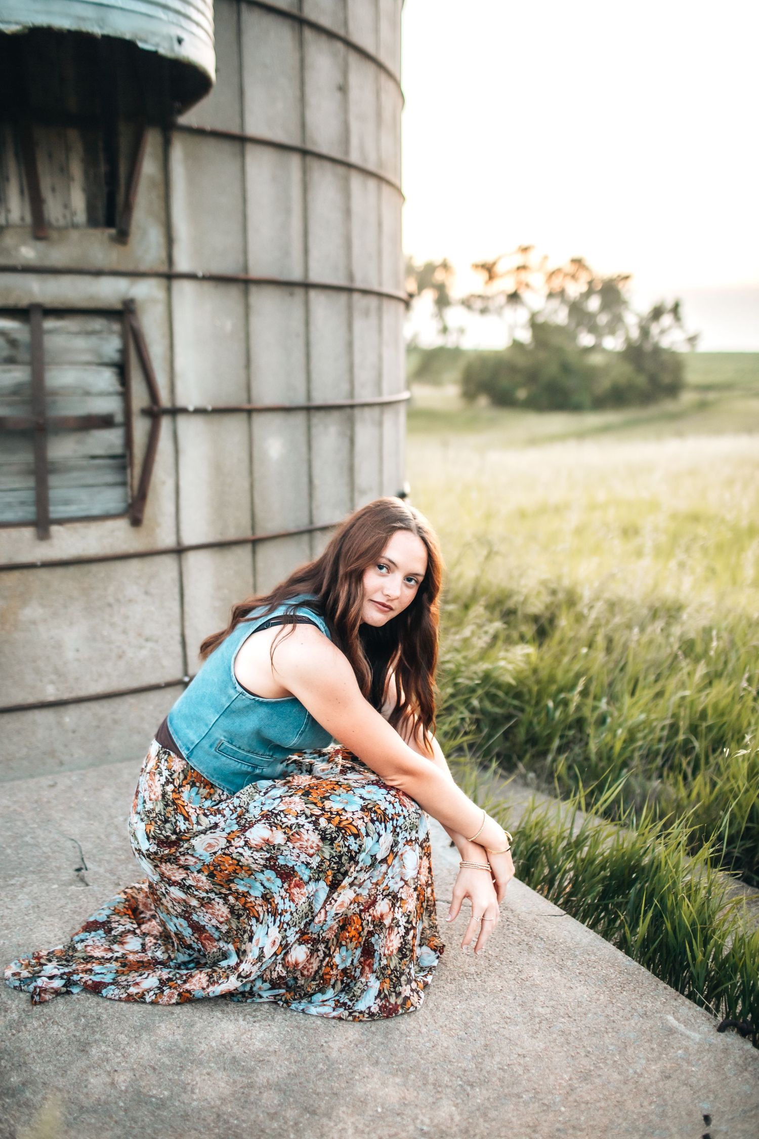 Young woman in floral maxi skirt and teal top posing beside a rustic grain silo in a green field at sunset.