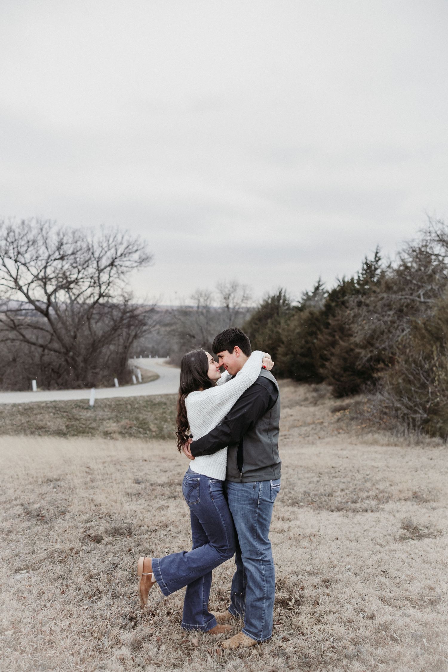 A couple embraces and almost kisses in a winter field with bare trees and a winding road in the background.