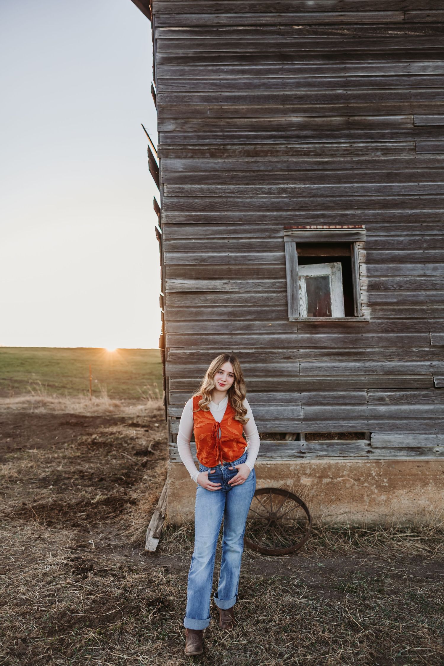 Young woman in orange vest and jeans poses by rustic wooden barn at golden sunset on rural farmland.