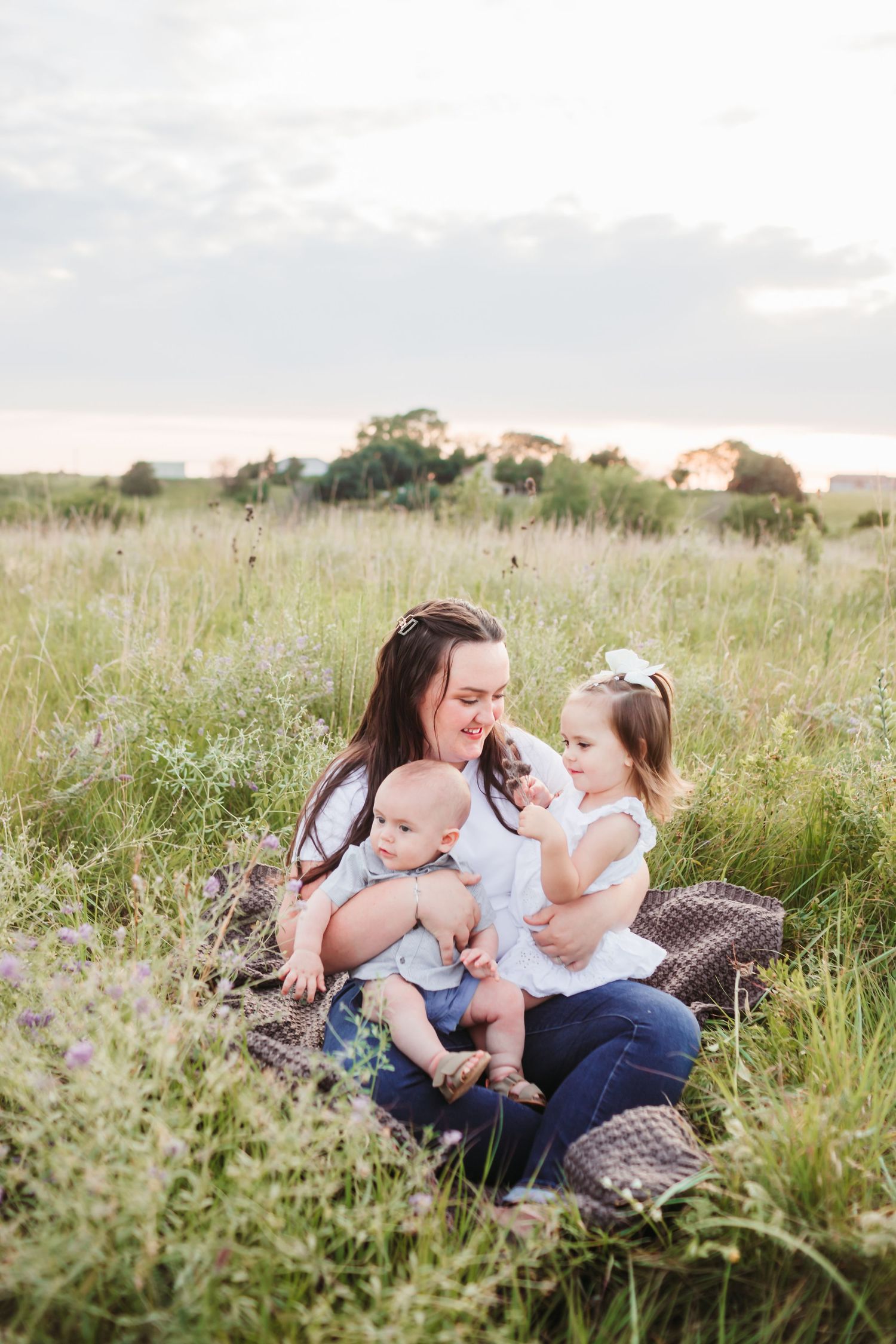 A tender mother and kid moment captured on a blanket in a sunlit meadow with tall grass and wildflowers.