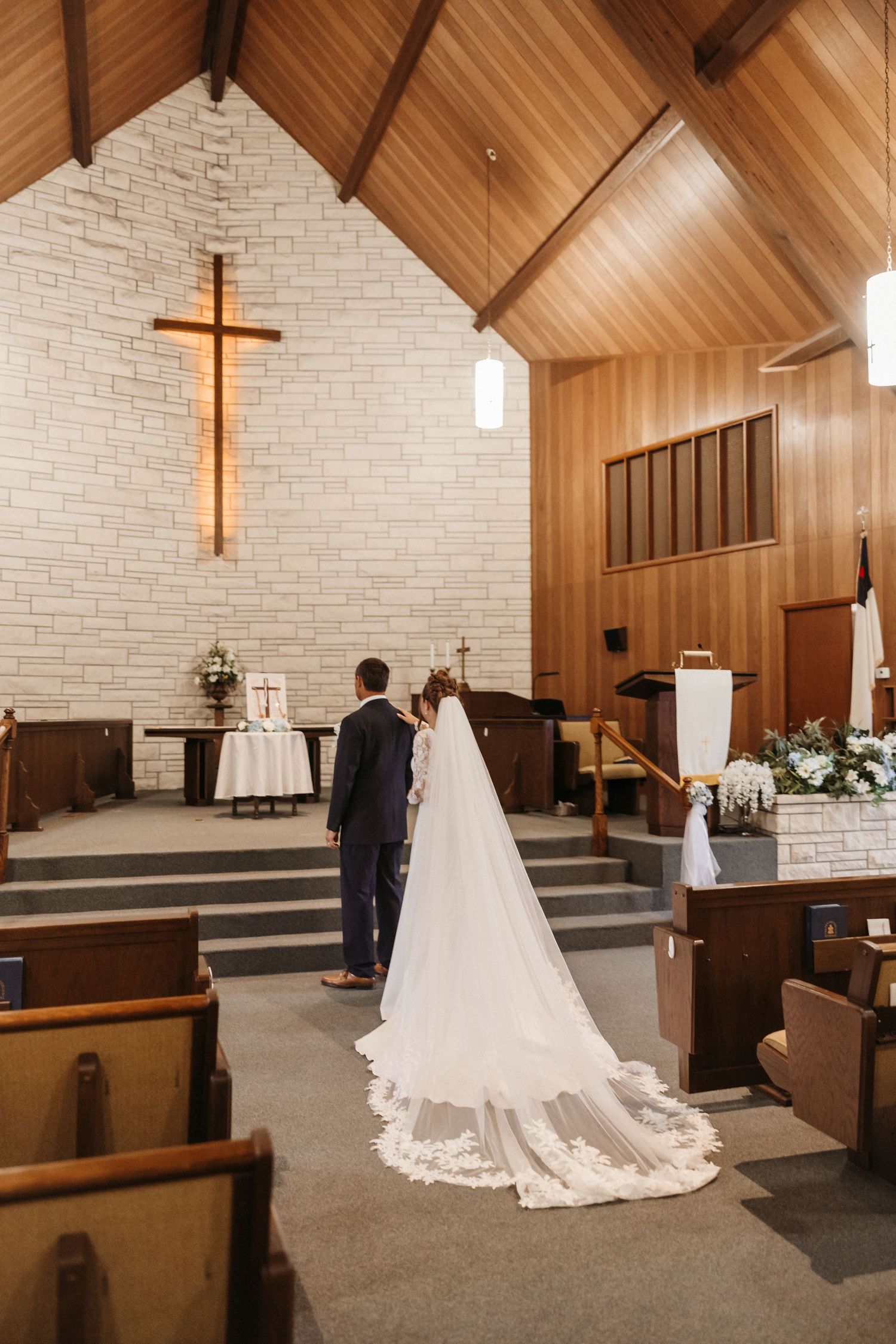 Bride and groom stand at church altar during wedding ceremony, bride wearing long lace veil trailing down aisle.